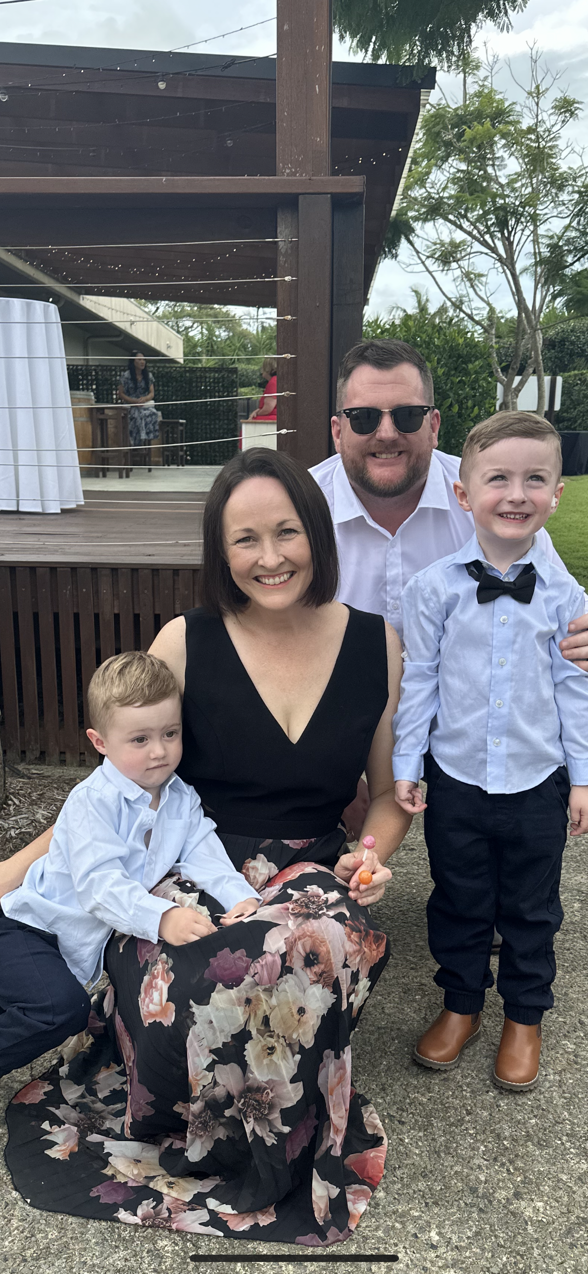 Group of four people at an outdoor event, smiling at the camera. A woman in a black floral dress kneels in front, holding a lollipop, with a young boy with blond hair and a white shirt to her left. Behind her are a man with sunglasses and a white shirt, and a young boy with a light blue shirt and bow tie standing to the right.