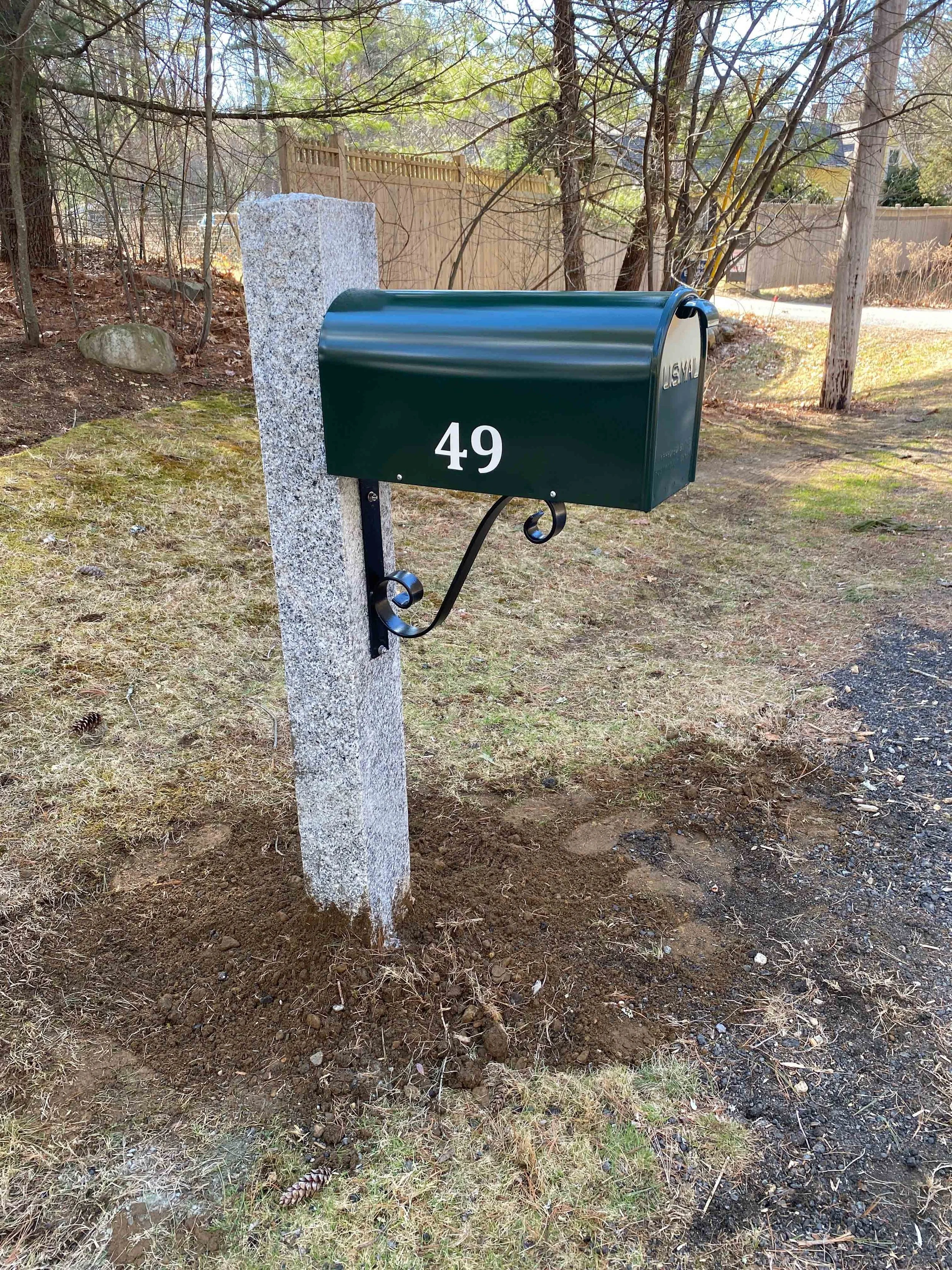 Green mailbox with the number 49 mounted on a stone post in a yard with grass, trees, and a wooden fence in the background.