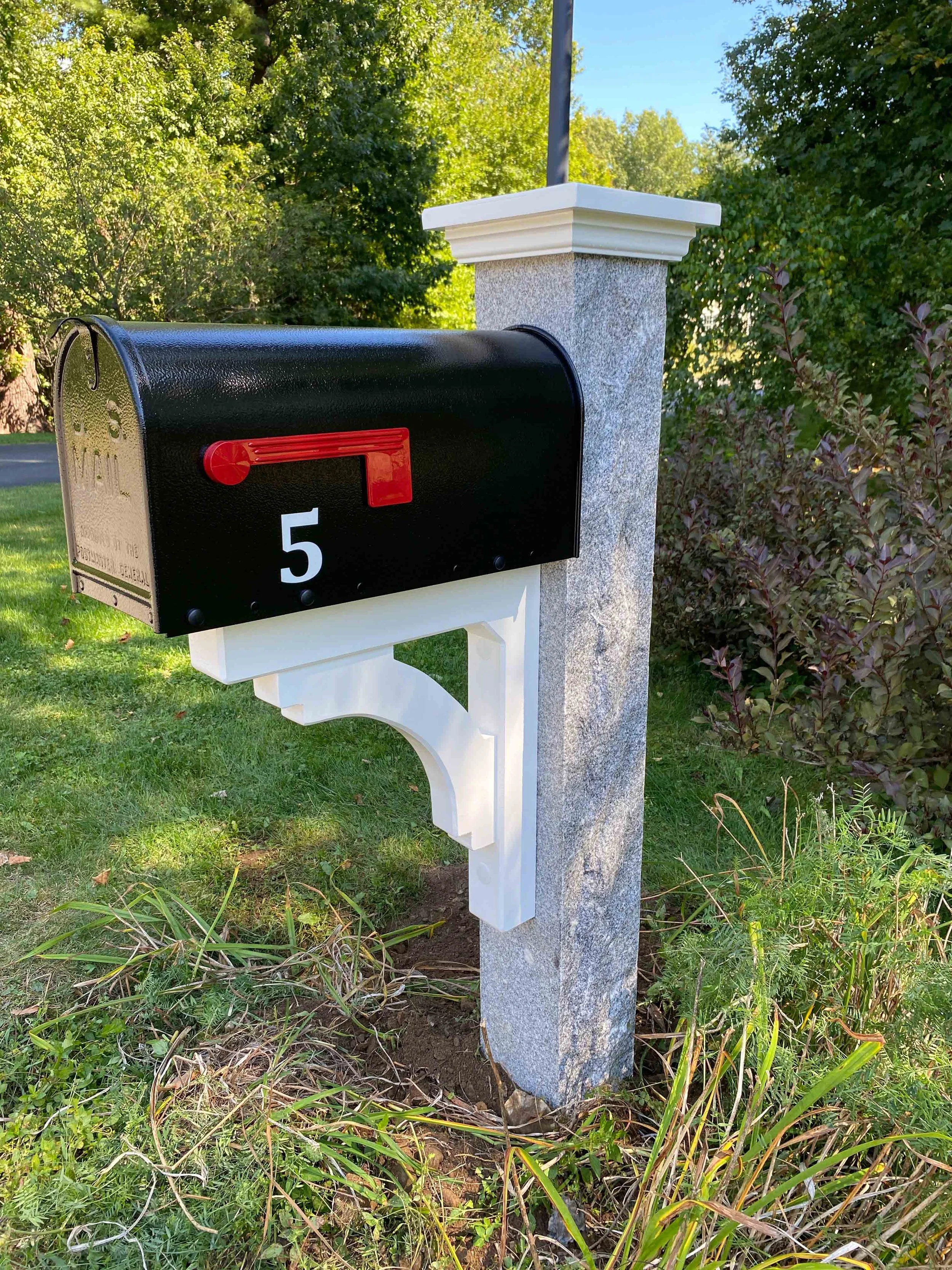 A black mailbox with a red flag and the number 5, mounted on a white decorative post next to a gray stone pillar, surrounded by green grass and bushes under a clear blue sky.