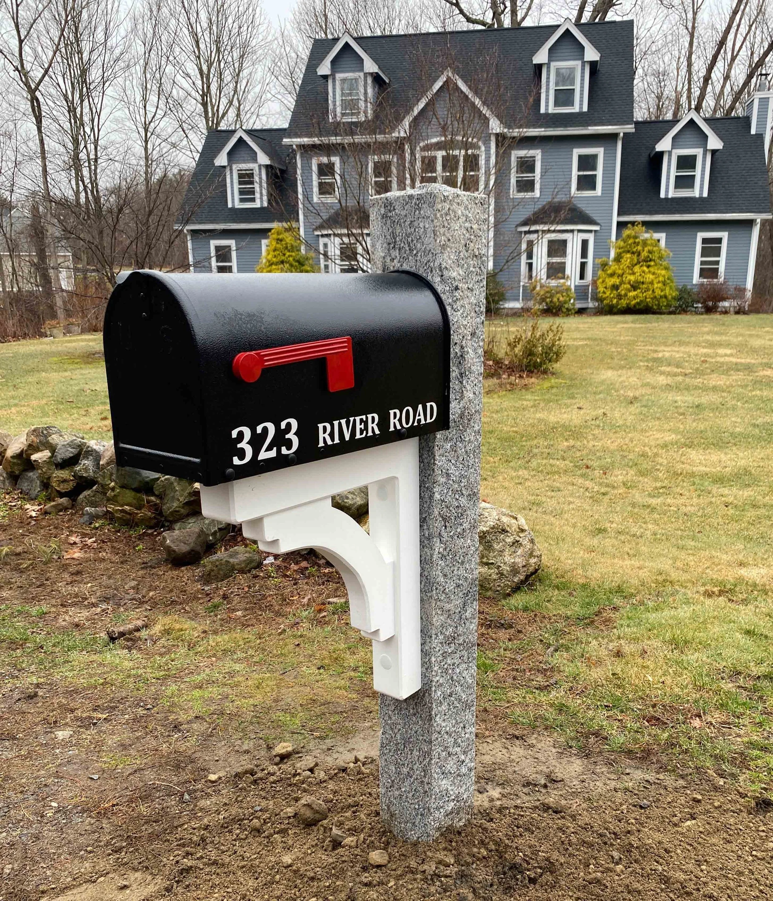 Black mailbox with red flag mounted on a white post, in front of a large black granite post, with a big house and yard in the background.