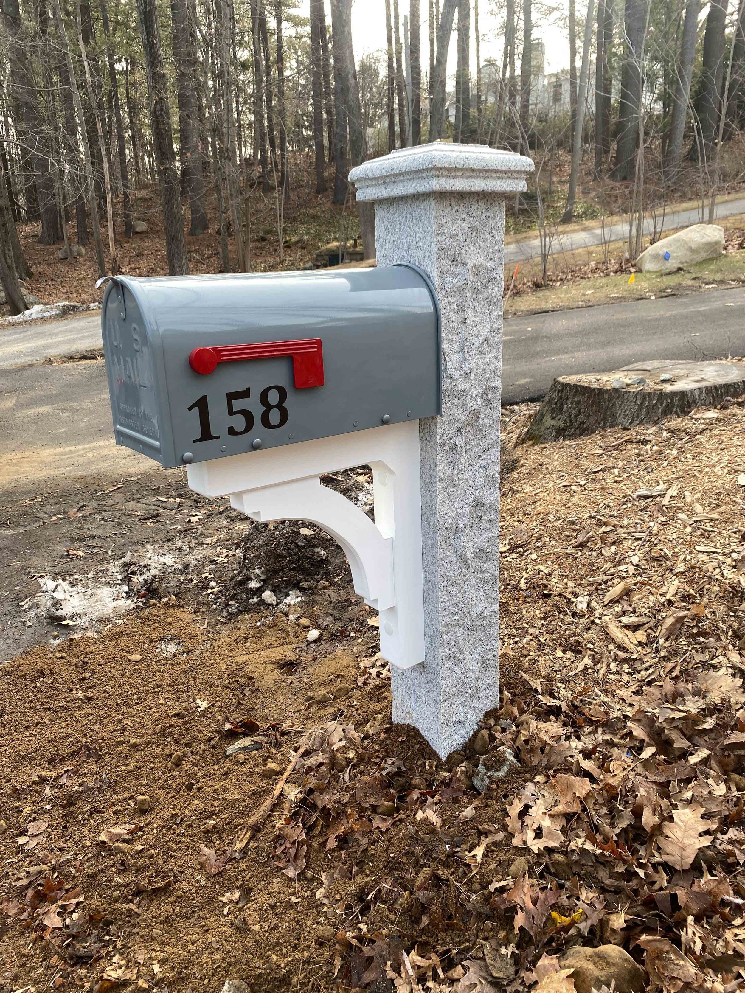 A grey mailbox with red flag displaying the number 158, mounted on a white post, next to a grey stone pillar on a dirt and leaf-covered ground in a wooded area.