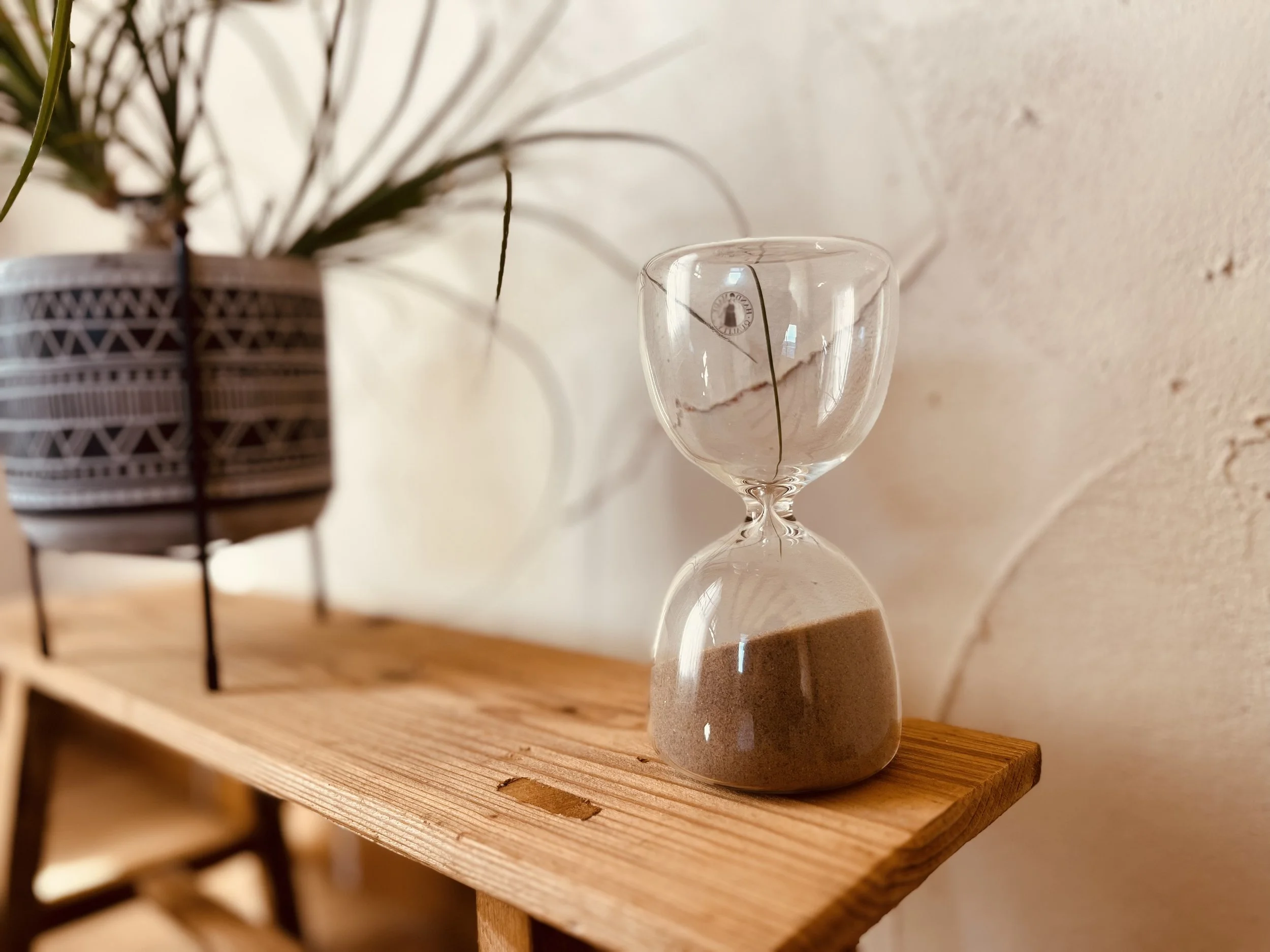 Un reloj de arena de vidrio con arena gris en una repisa de madera, junto a una planta decorativa en una maceta con patrones geométricos.