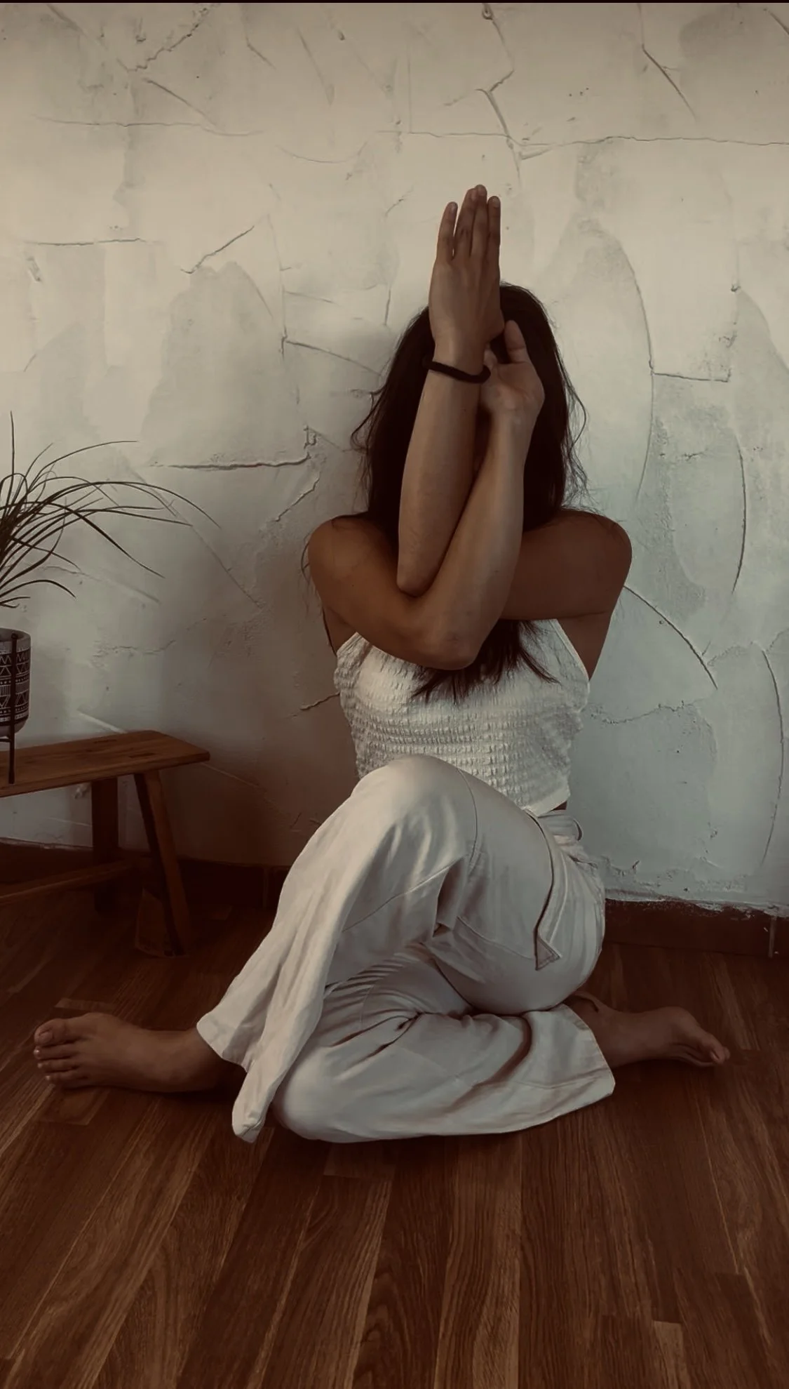 Mujer en posición de yoga en un piso de madera, con pared de fondo, posando con brazos cruzados y cubriéndose la cara.