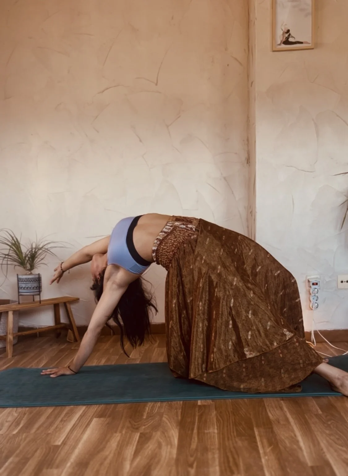 Mujer haciendo yoga en una habitación con pared de textura suave y piso de madera, en posición de gato de yoga, con una planta en la mesa y un cuadro en la pared.