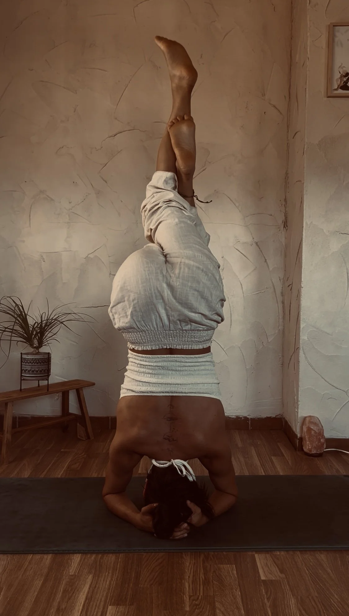 Persona haciendo la postura de equilibrista en yoga, en posición invertida con cabeza en el suelo y piernas extendidas hacia arriba, en una habitación con paredes de color crema y piso de madera.