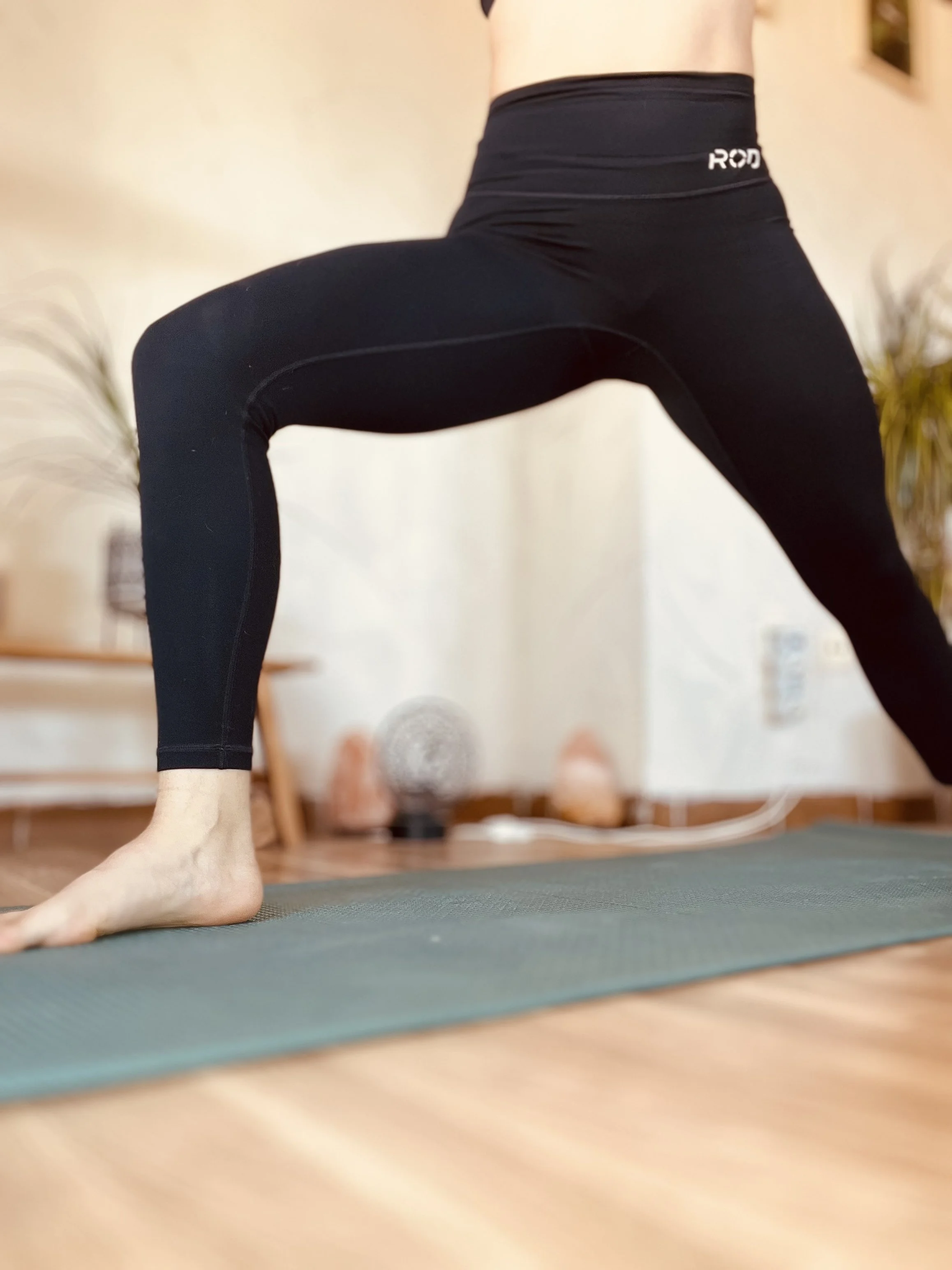 Persona practicando yoga en postura de la expansiva o guerrero en un ambiente interior. Utiliza ropa deportiva negra y está sobre una colchoneta de yoga.