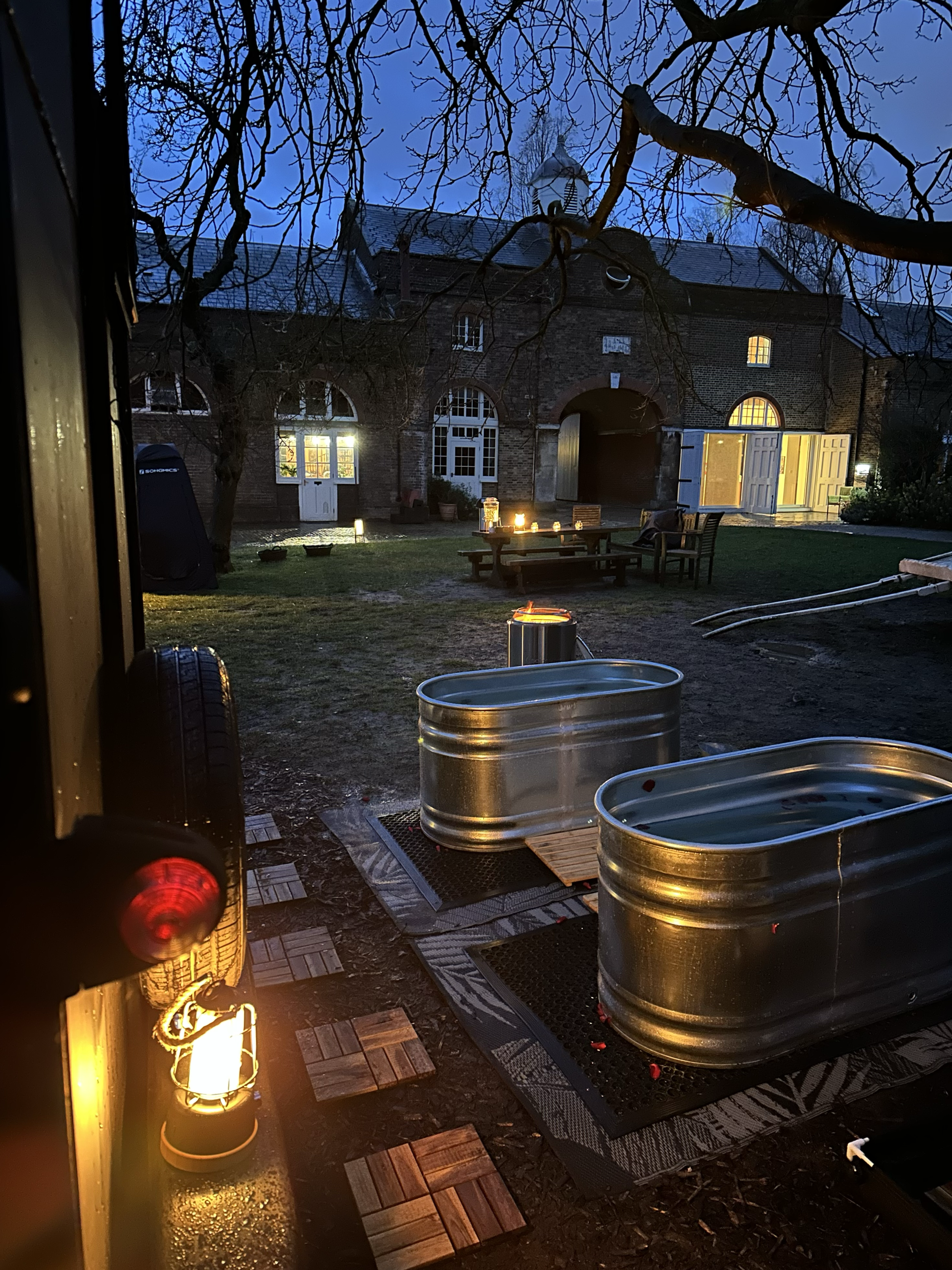A stable yard scene at dusk with two small metal tubs, a wooden deck, outdoor lanterns, and a picnic table with lit candles. In the background, a brick building with large windows and open doors, and leafless trees are visible.