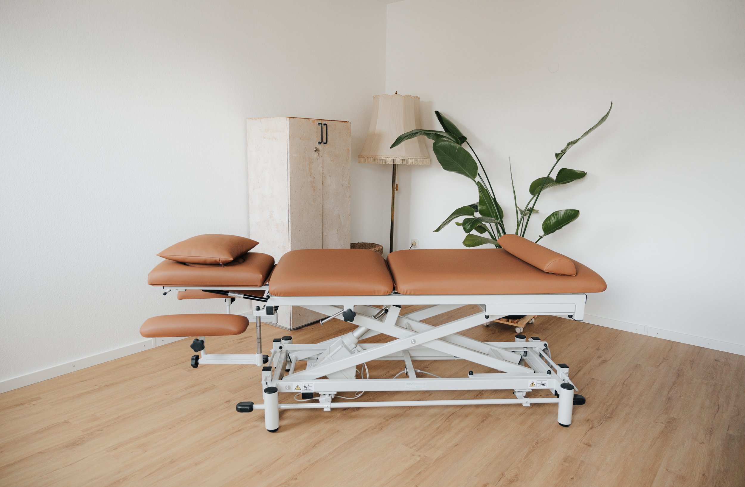 Chirurgischer Behandlungstisch in einem Behandlungsraum, mit braunen Polstern, neben einer Stehlampe, einer Pflanze und einem weißen Schrank, auf Holzfußboden.