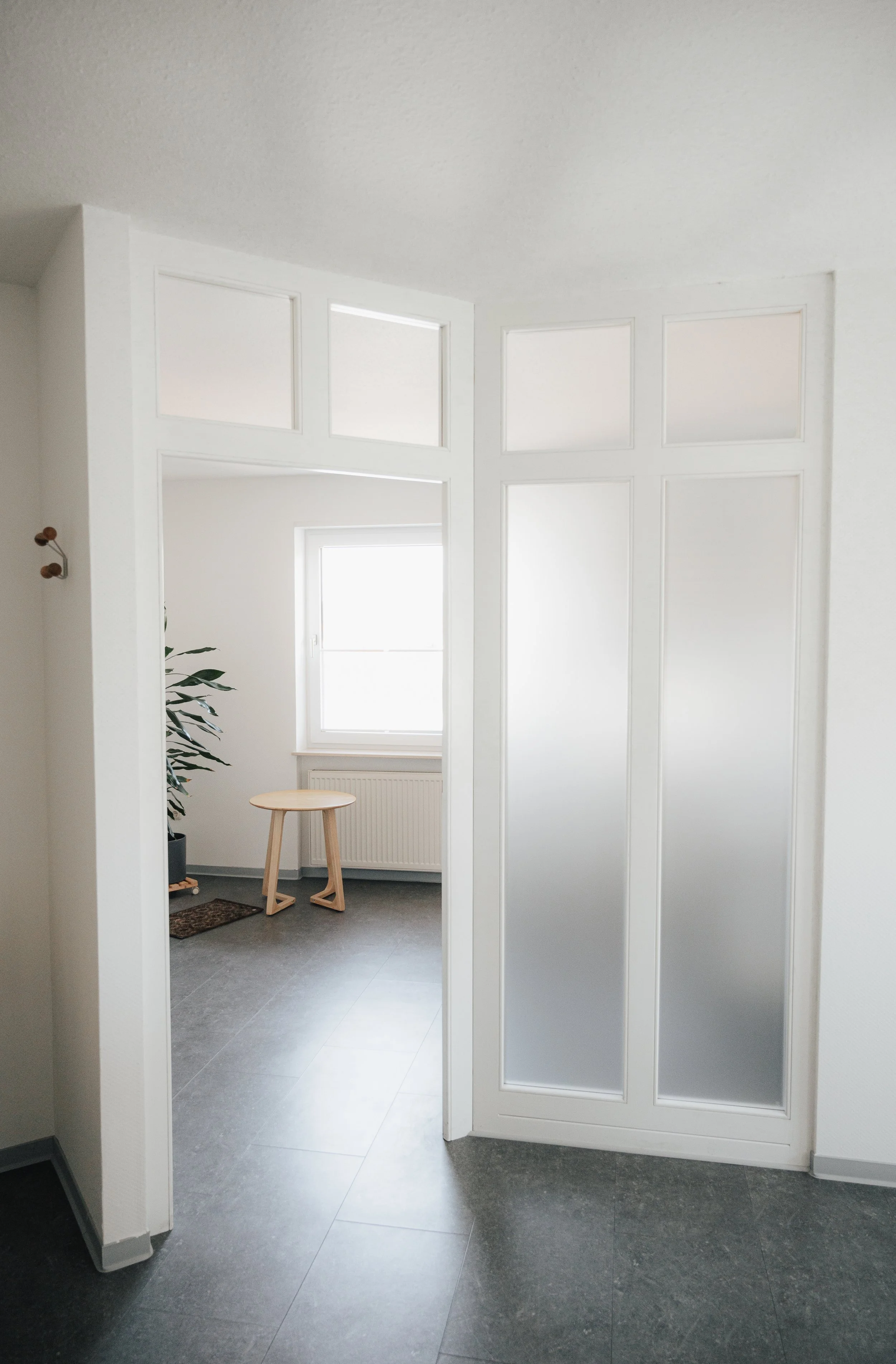 Ein helles, modernes Raum mit weißen Wänden, einem Fenster und einer Glastür. Es gibt einen kleinen Holztisch, eine Zimmerpflanze und eine Heizkörper unter dem Fenster.