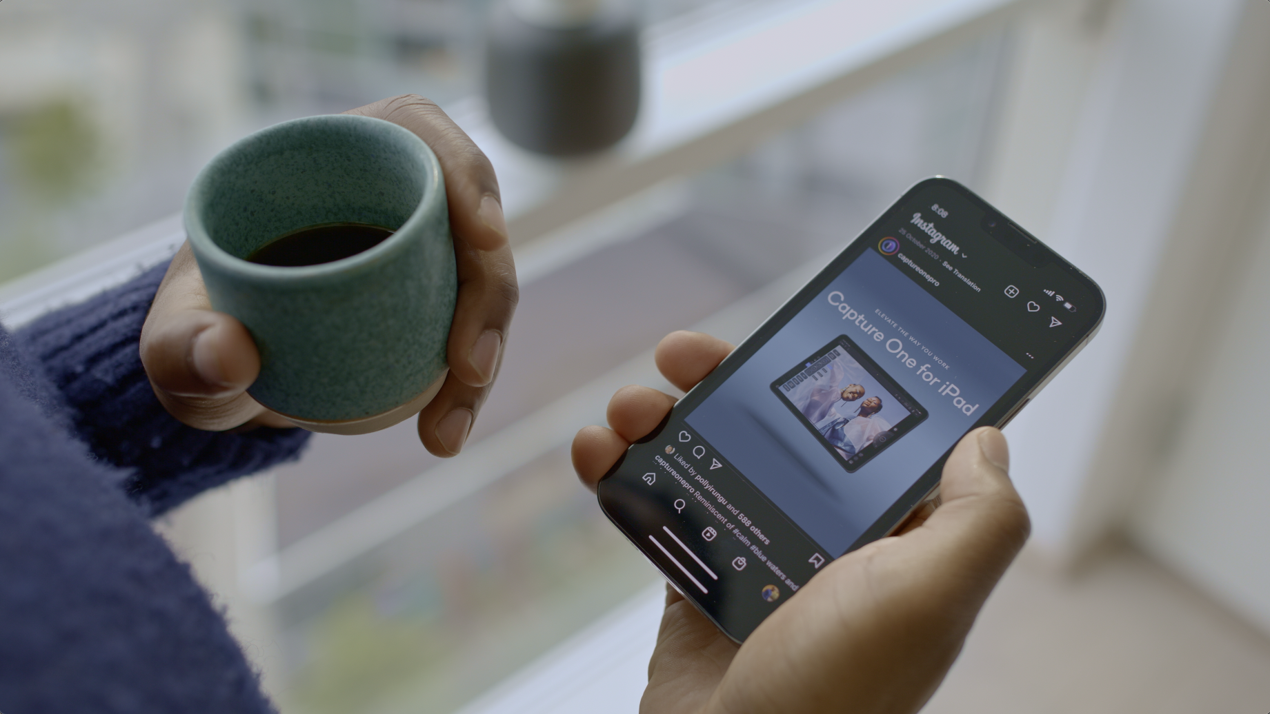 Person holding a coffee cup in one hand and a smartphone displaying an Instagram post about the Capture One app in the other hand.
