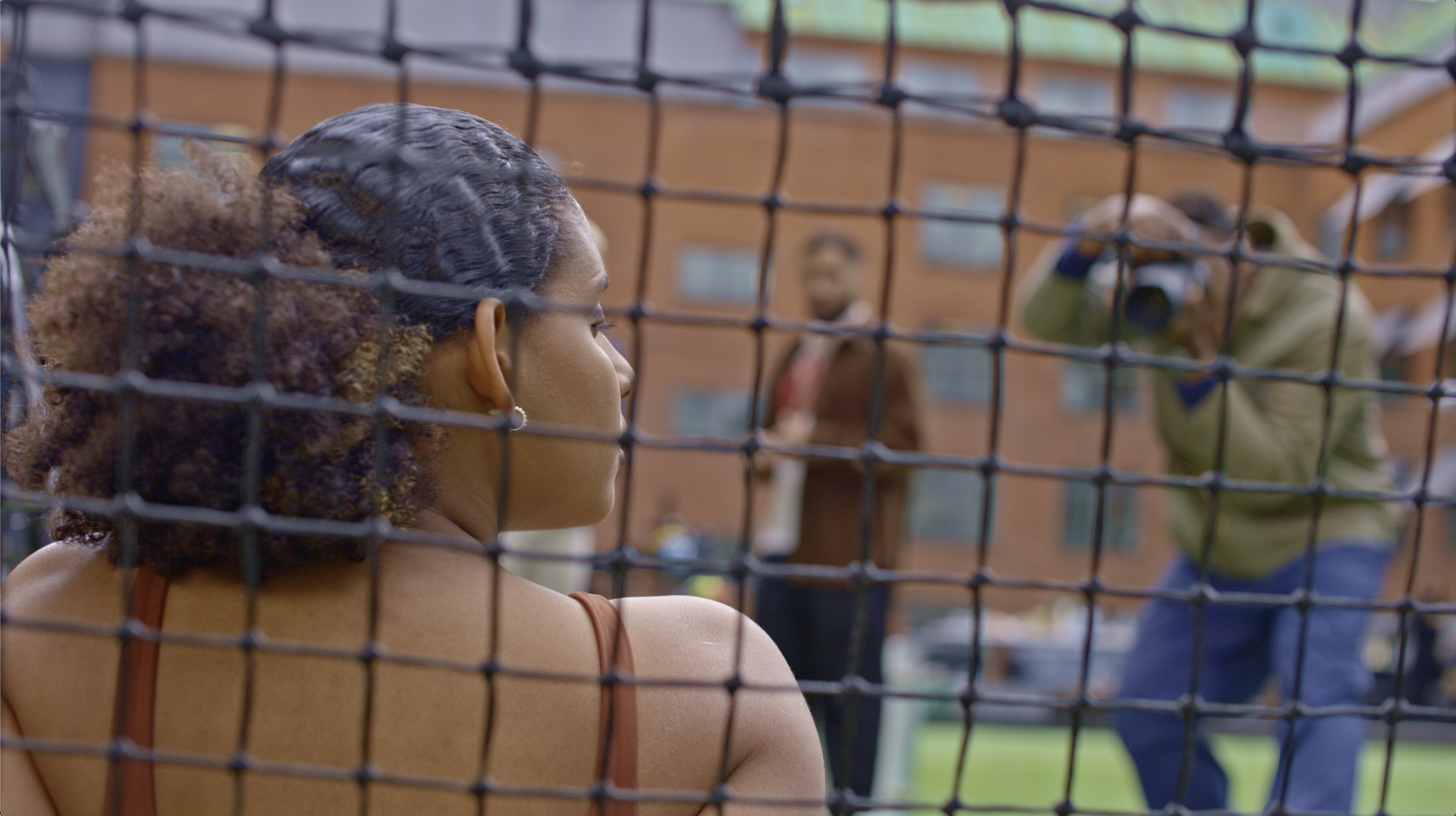 A woman with curly hair looking through a tennis net at two men on the other side, one taking a photo and the other adjusting his hat, with a building in the background.