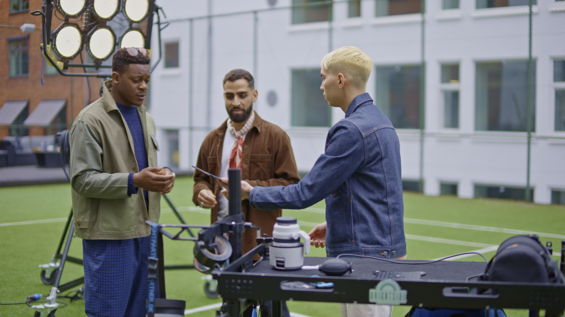 Three men stand on a sports field with equipment, discussing a tablet, with film lighting and tech devices nearby.