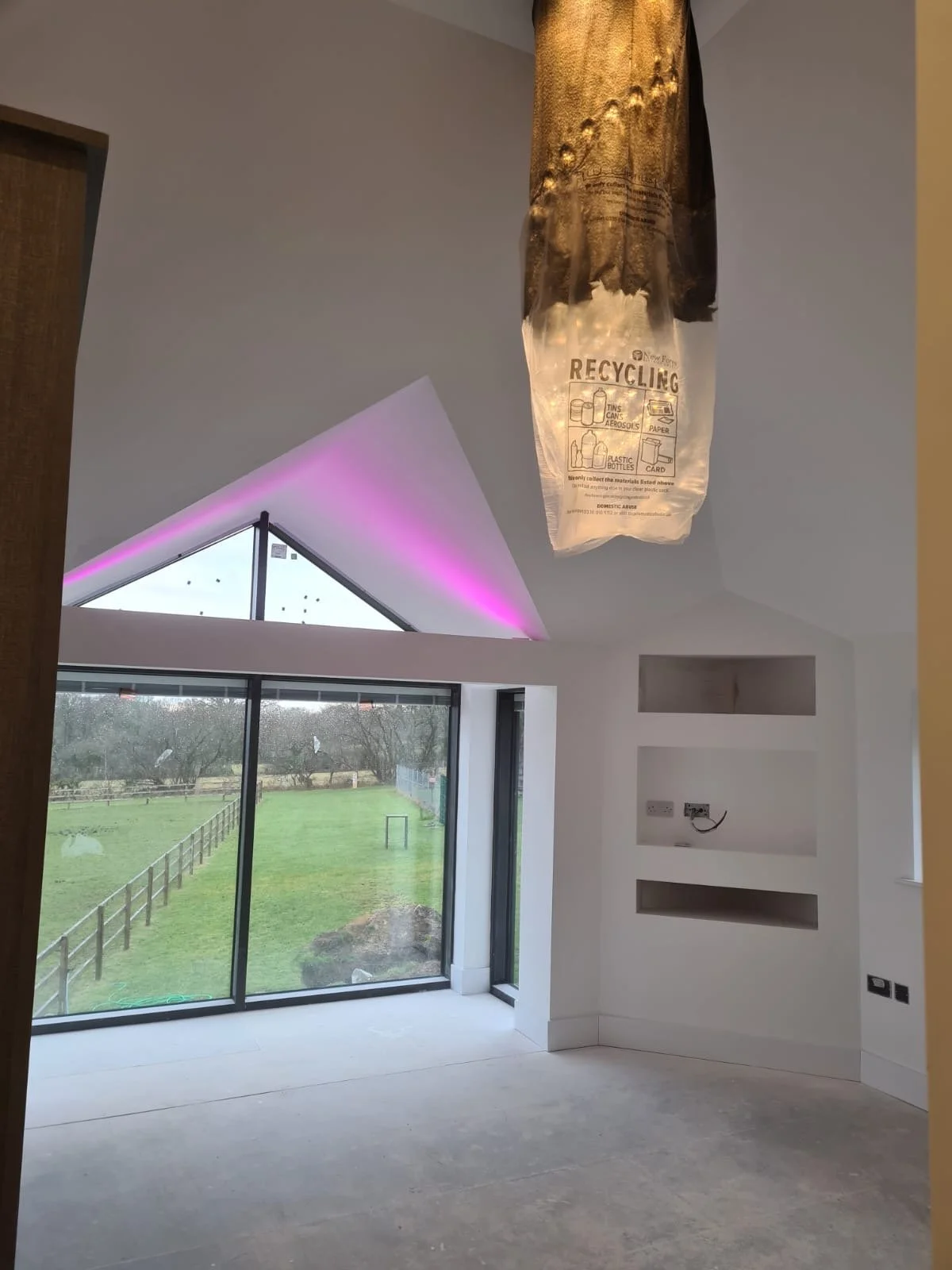 An unfinished room with a large window showing a lawn outside, recessed shelves in the wall, and a ceiling light fixture made from a recycled plastic shopping bag.