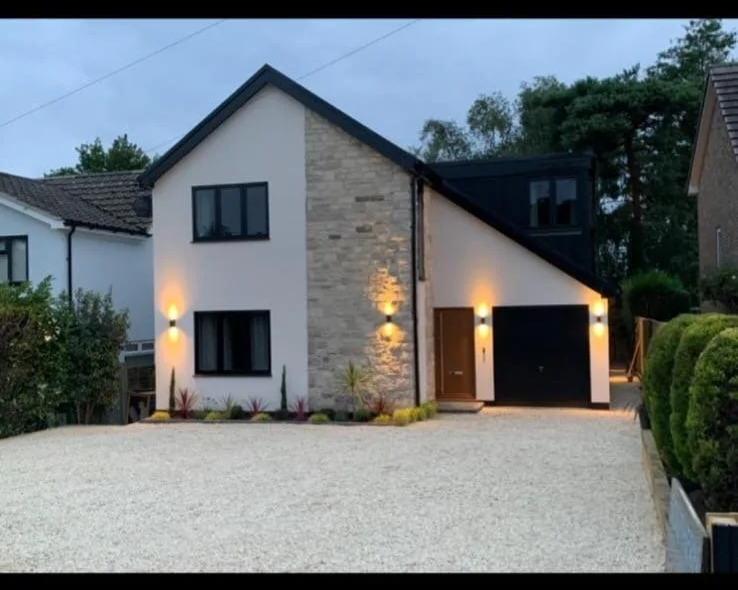 Modern two-story house with a stone and white exterior, black windows, a black garage door, and exterior wall-mounted lights, surrounded by a gravel driveway and landscaped plants.