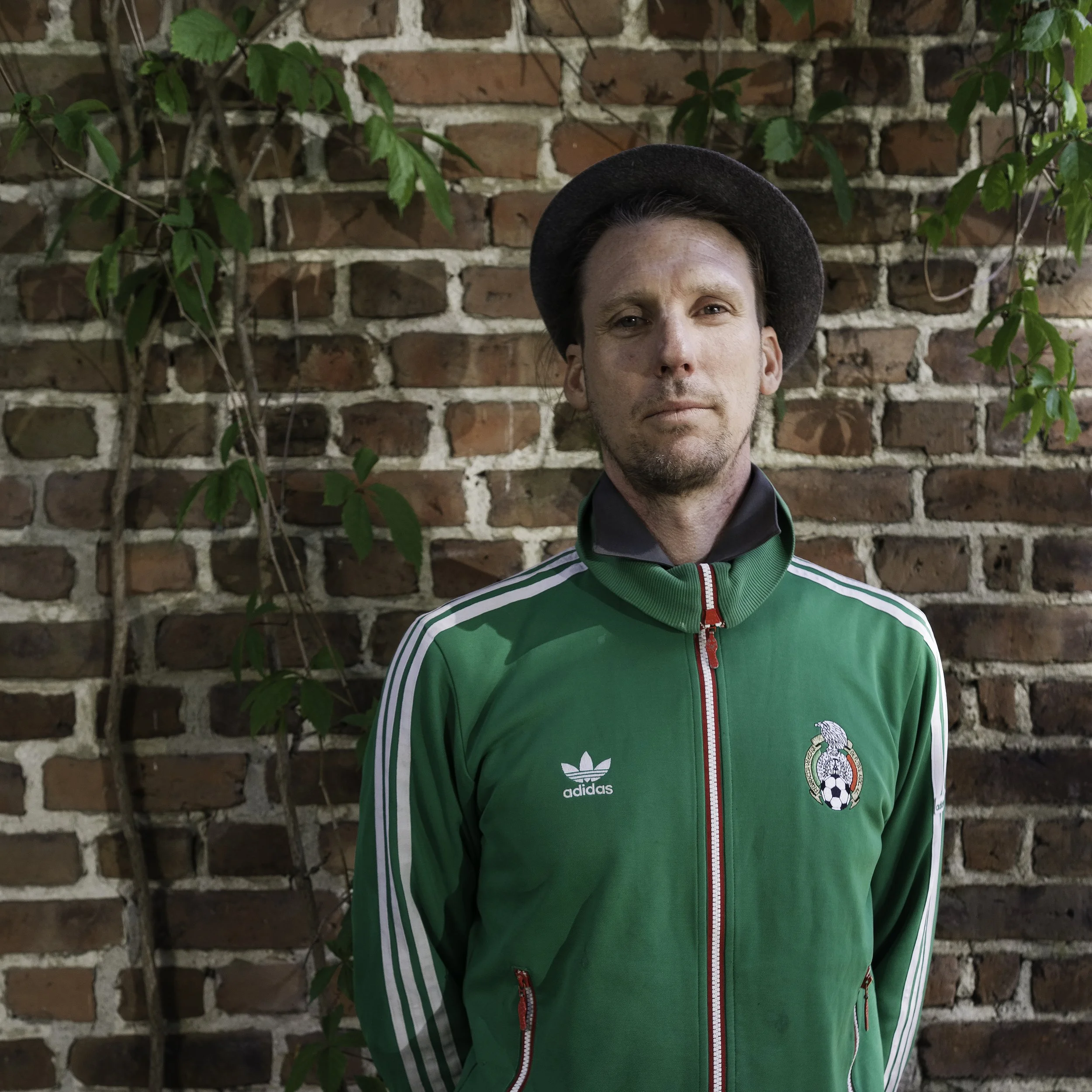 Et bilde av en mann som står foran en murvegg med grønt løv. Mannen har på seg en grønn Adidas-jakke og en svart hatt. Han har lyst hår og ser direkte inn i kameraet.