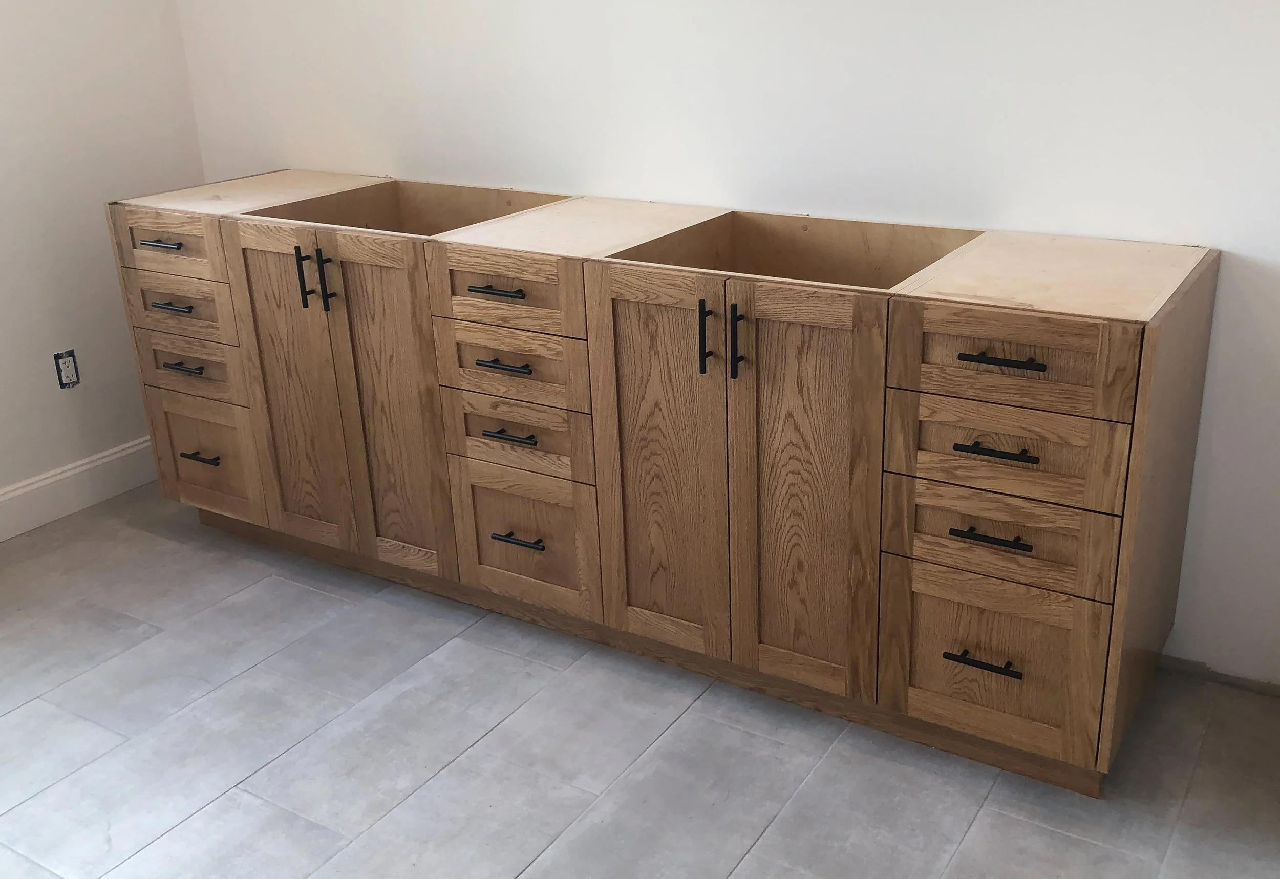 Wooden kitchen cabinet with multiple drawers and doors, partially built, in a room with gray tiled flooring and a plain beige wall.