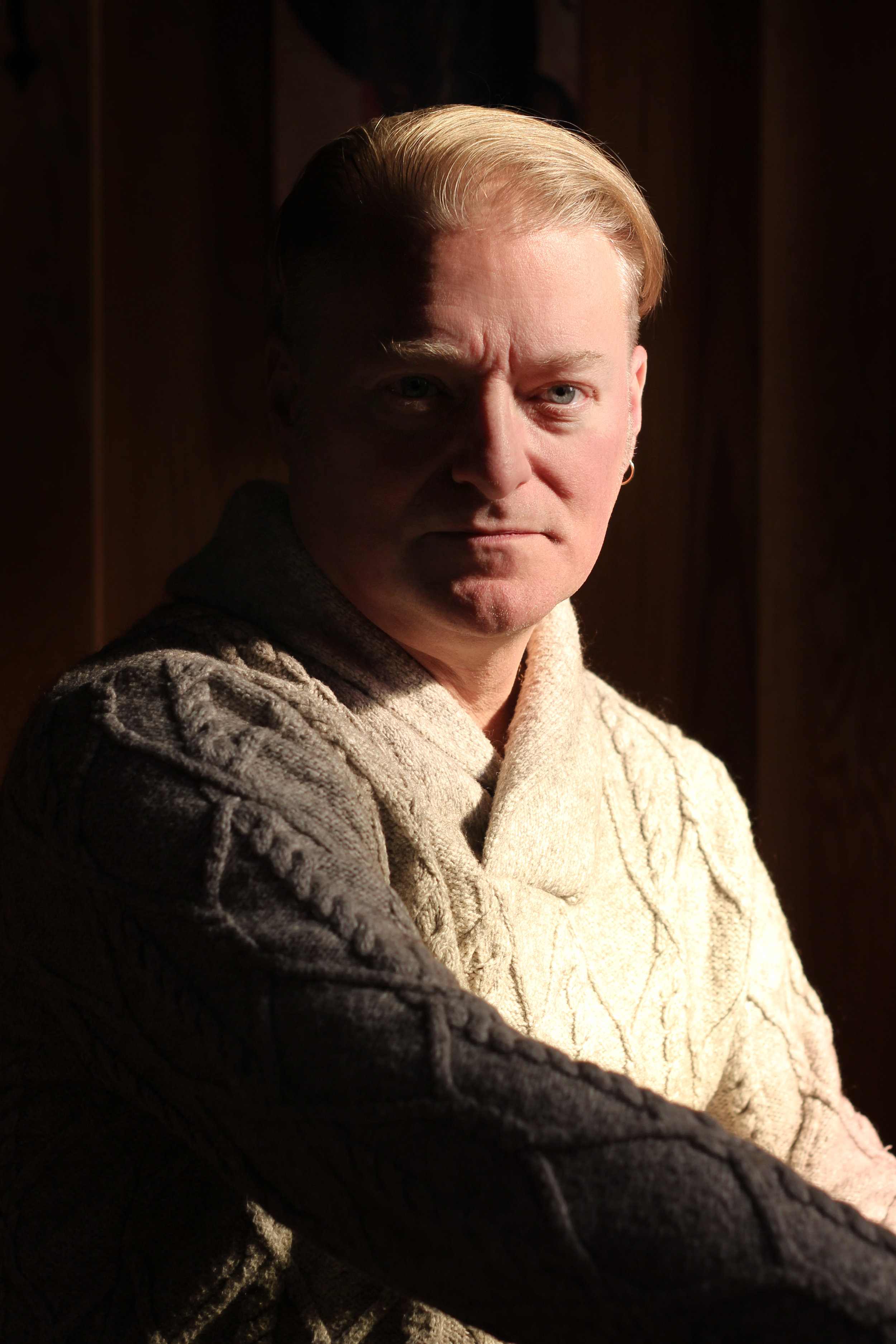A man with blonde hair wearing a beige and gray cable knit sweater, sitting against a dark wooden background with dramatic lighting.