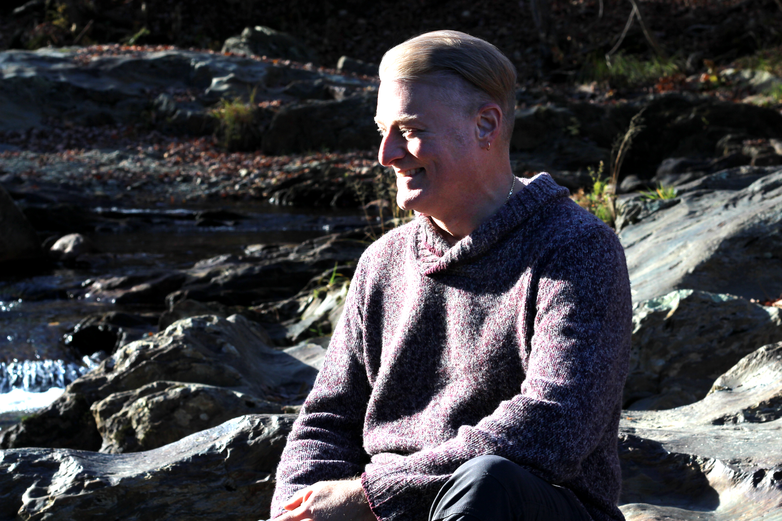 A person with short blond hair wearing a gray sweater sitting outdoors near a rocky stream, smiling and enjoying nature.
