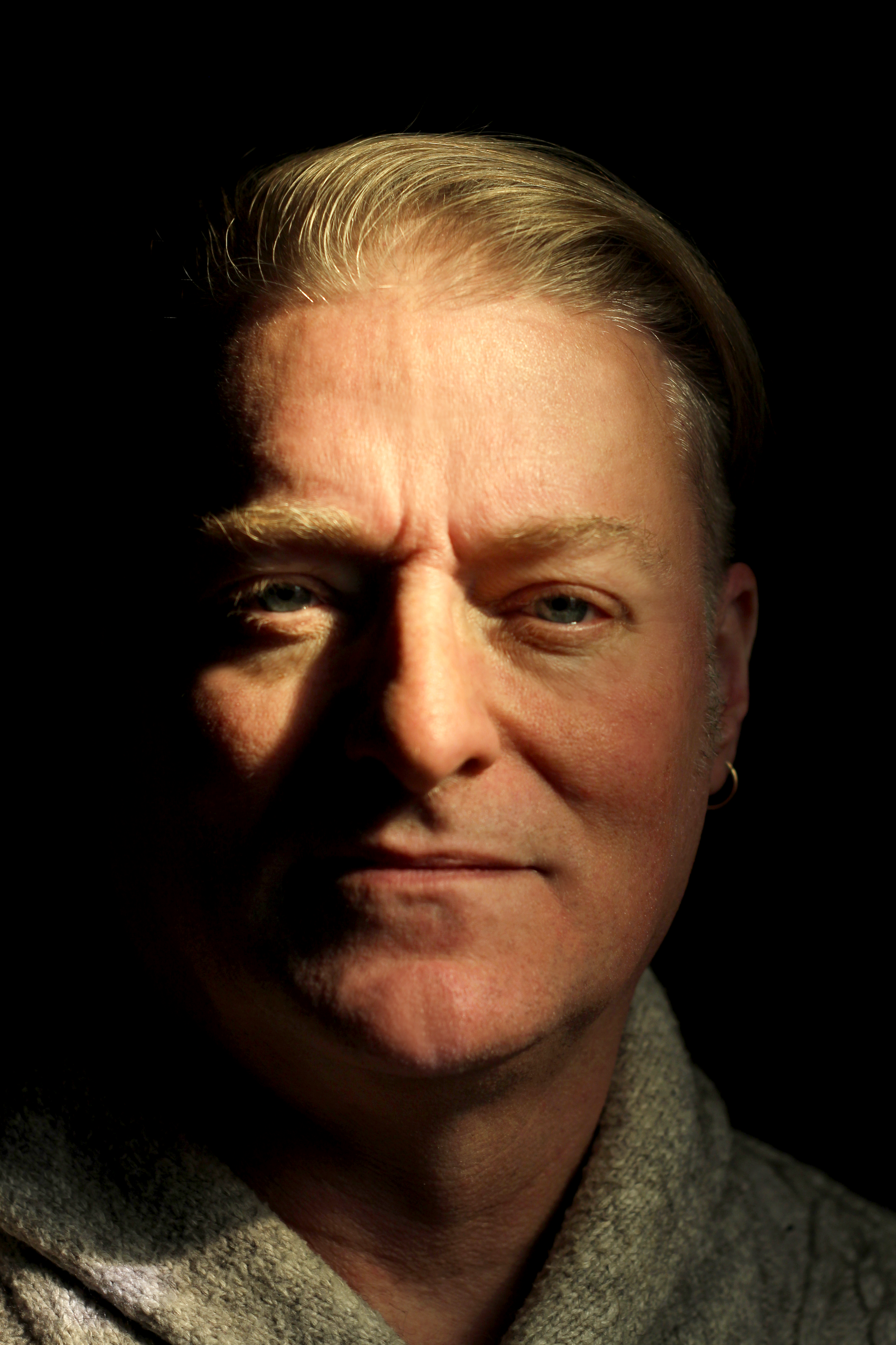 Close-up of a man's face with partial shadows, light on the right side, showing blonde hair, light eyes, and wearing a gray sweater.