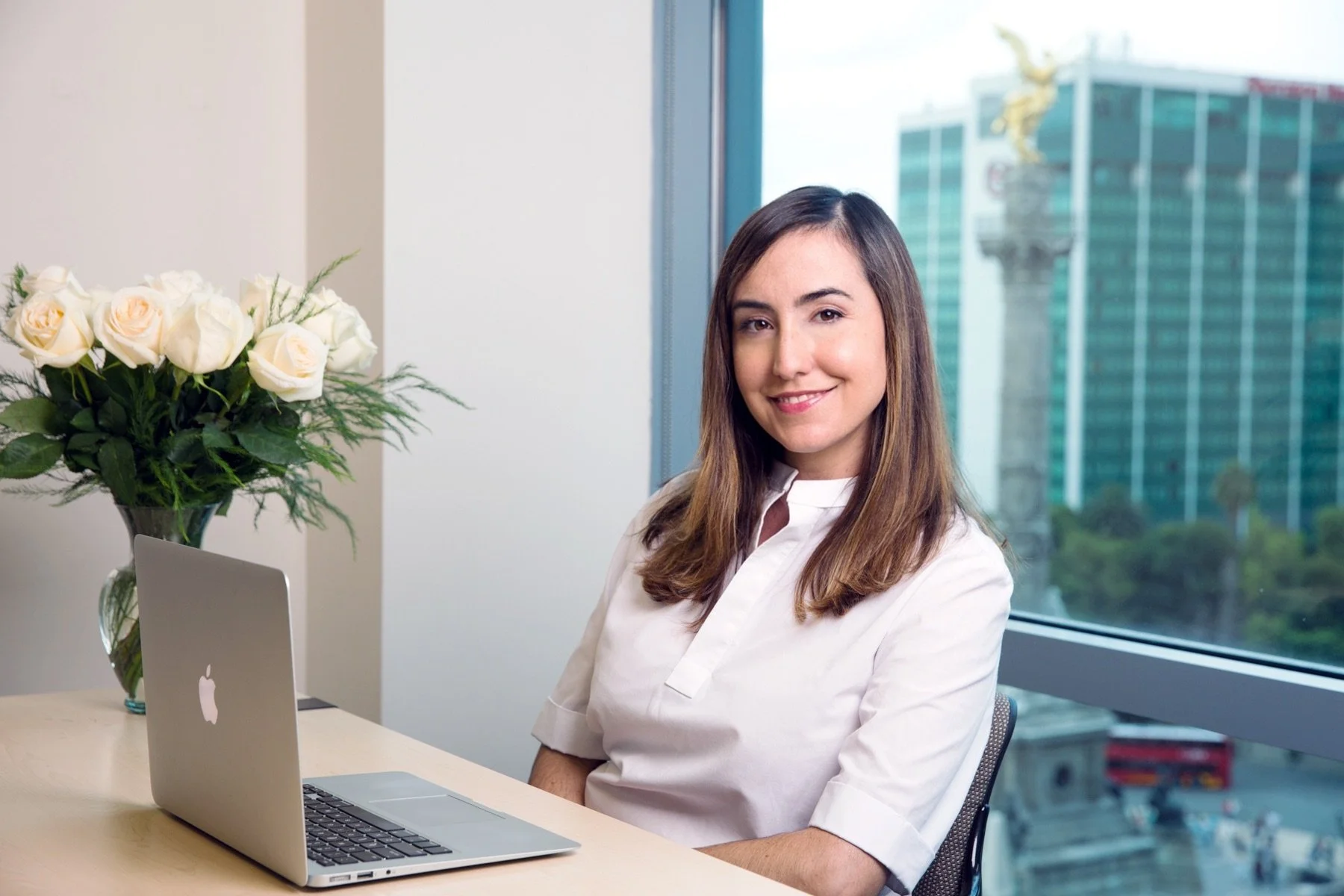 A woman sitting at a desk with a laptop and a vase of white roses near a window showing city buildings outside.