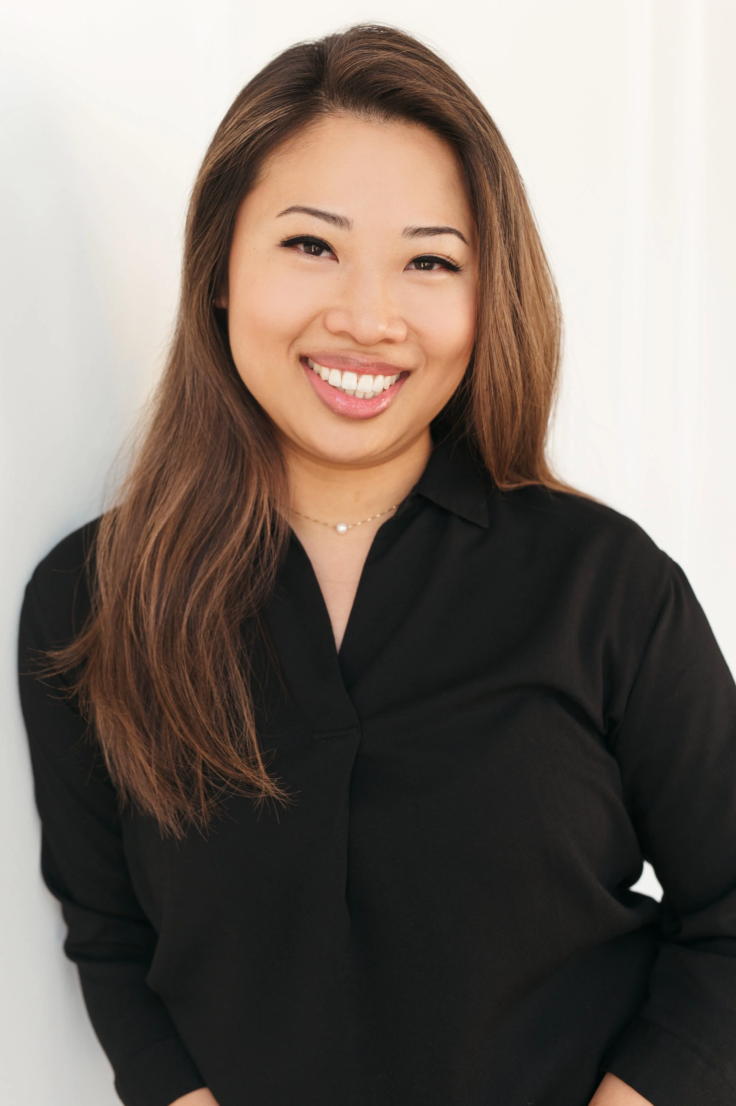 A woman with long brown hair, wearing a black top, smiling, with a white background.