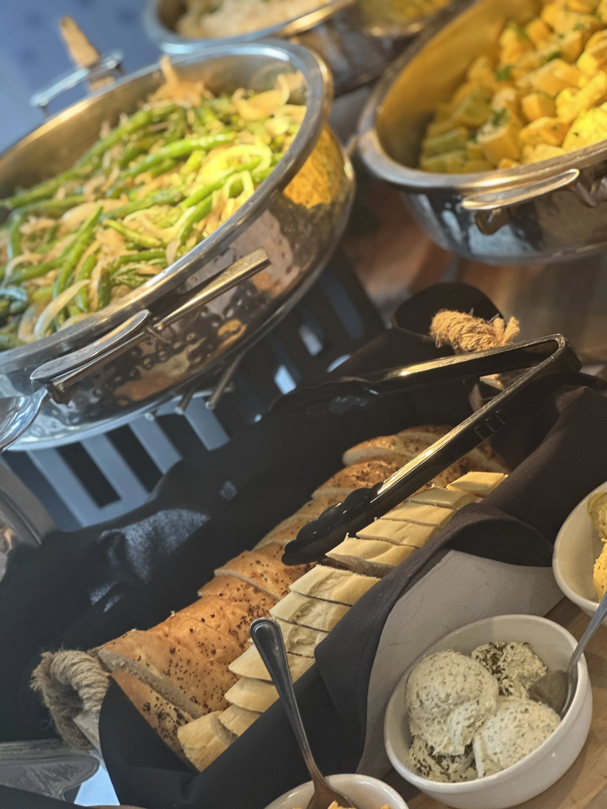 Buffet table with sliced bread, bowls of herbs and herbs, and containers of salad and vegetables.