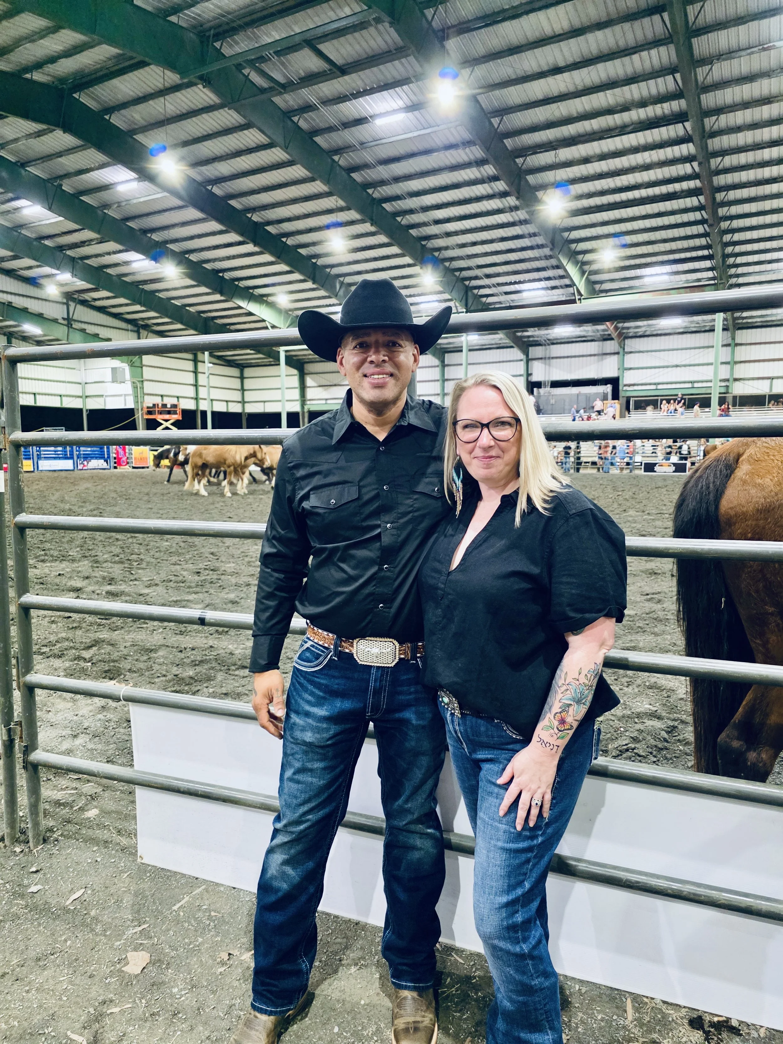 A man and woman standing inside an indoor rodeo arena, posing for a photo. The man is wearing a black cowboy hat, black long-sleeve shirt, jeans, and cowboy boots. The woman is wearing glasses, a black shirt, jeans, and has a tattoo on her arm. There are fans and other people in the background, along with a horse on the right side of the image.