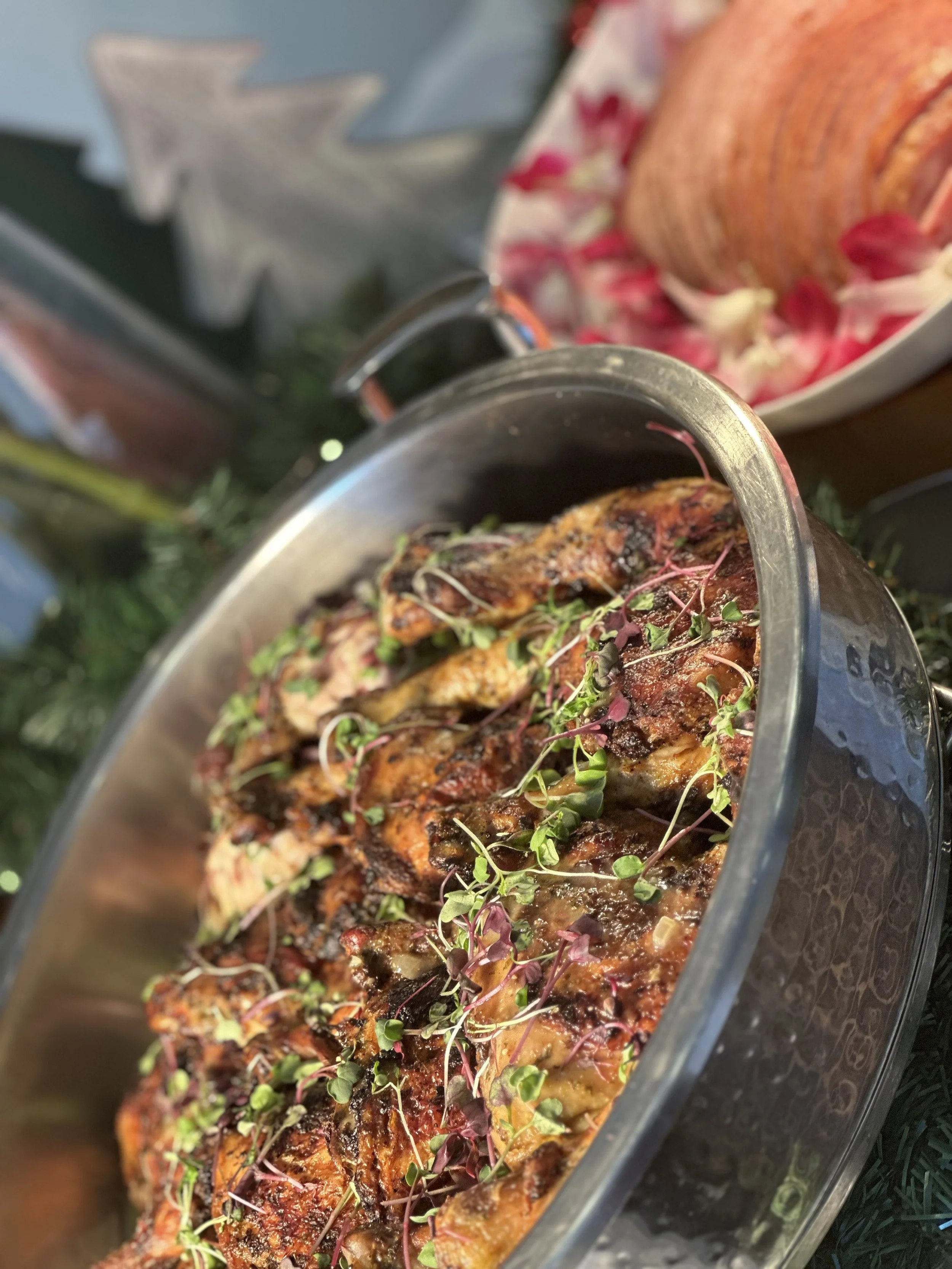 A roasted chicken garnished with microgreens on a serving dish, with a side dish of ham in the background. The food is presented on a table at a festive or holiday meal.