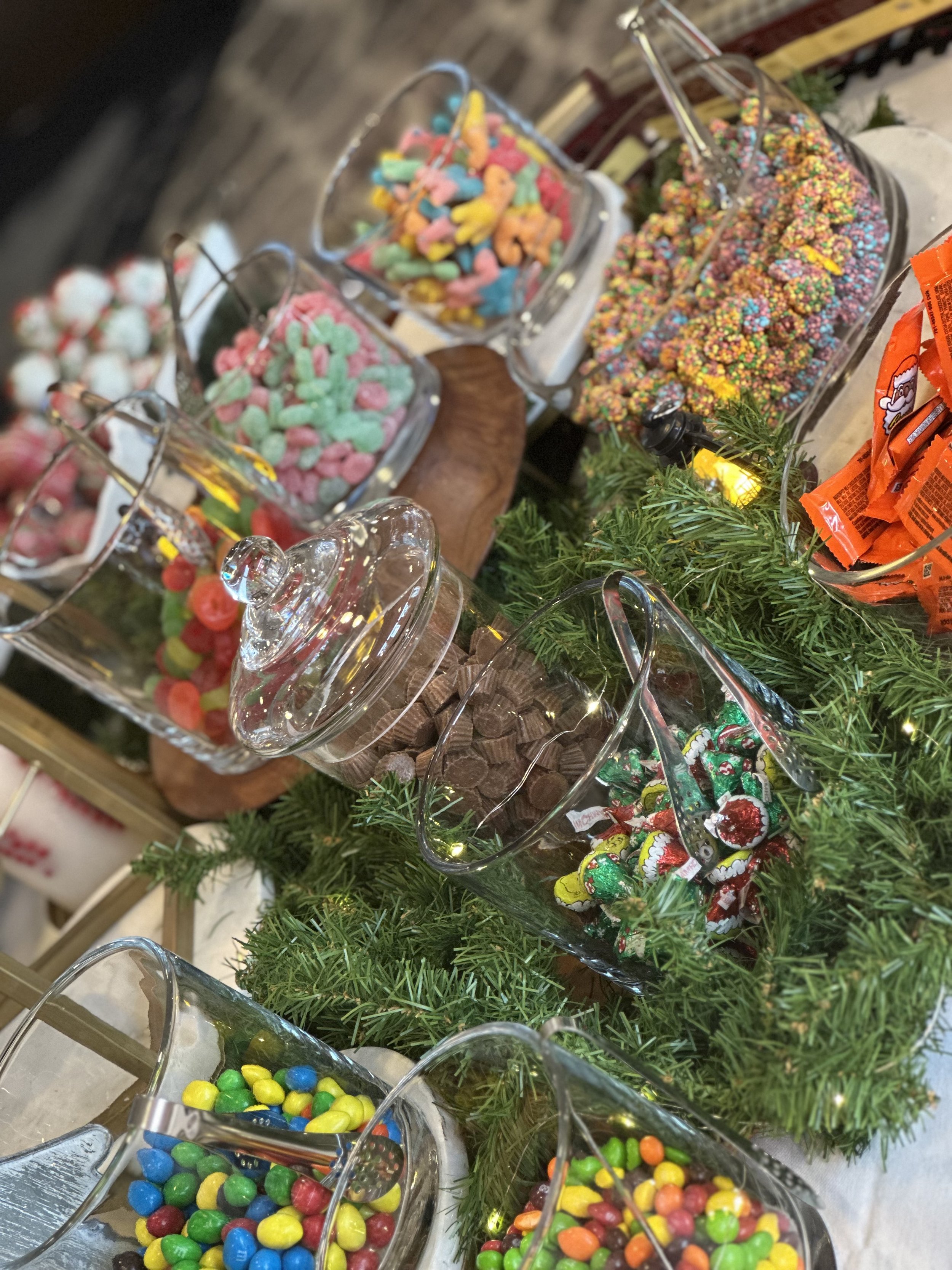 Assorted colorful candies in glass jars displayed on a table decorated with green artificial pine branches.
