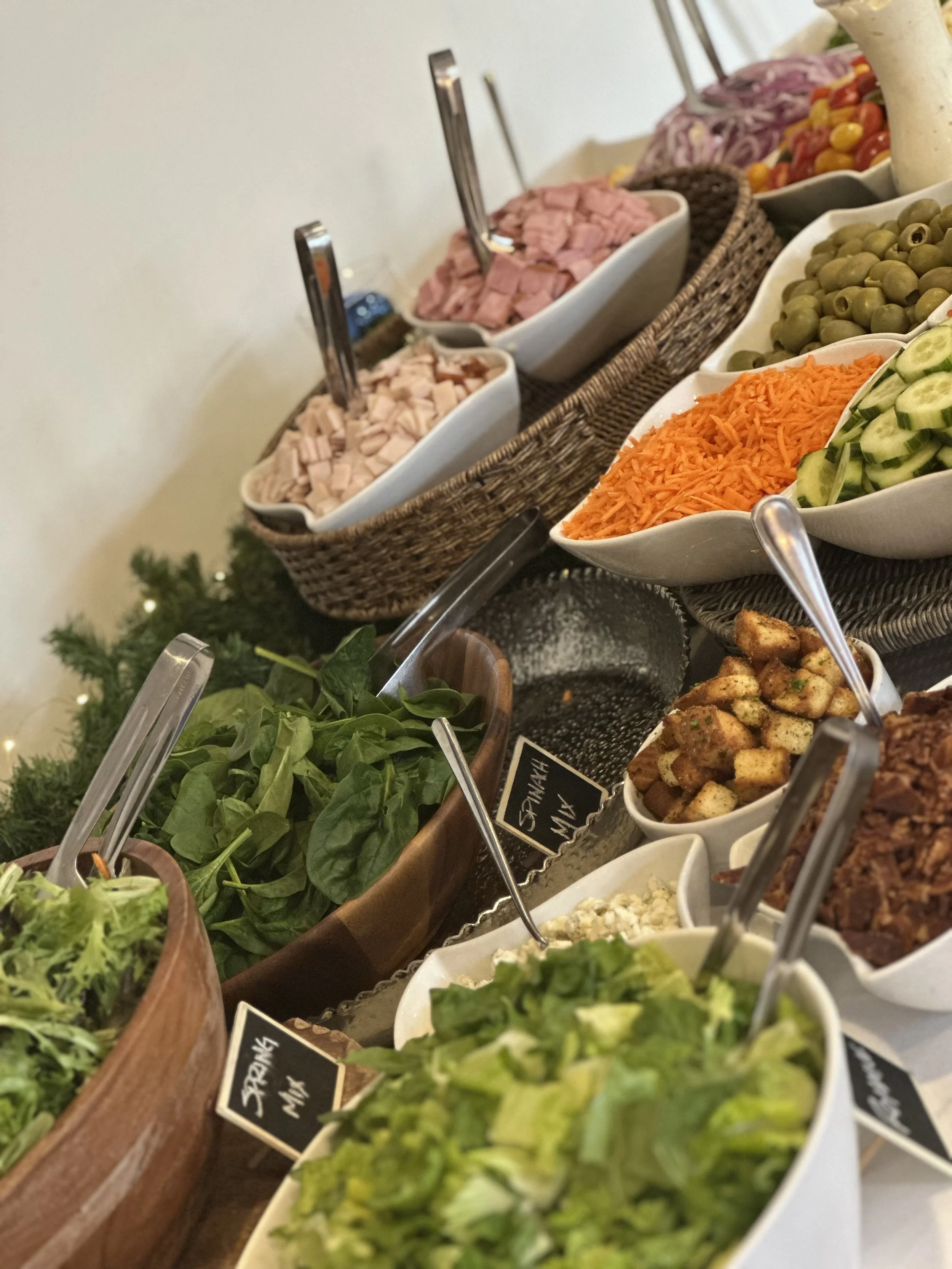 A salad bar with bowls containing various vegetables, meats, and toppings, including cherry tomatoes, shredded carrots, cucumbers, green olives, chopped ham, and fresh leafy greens.