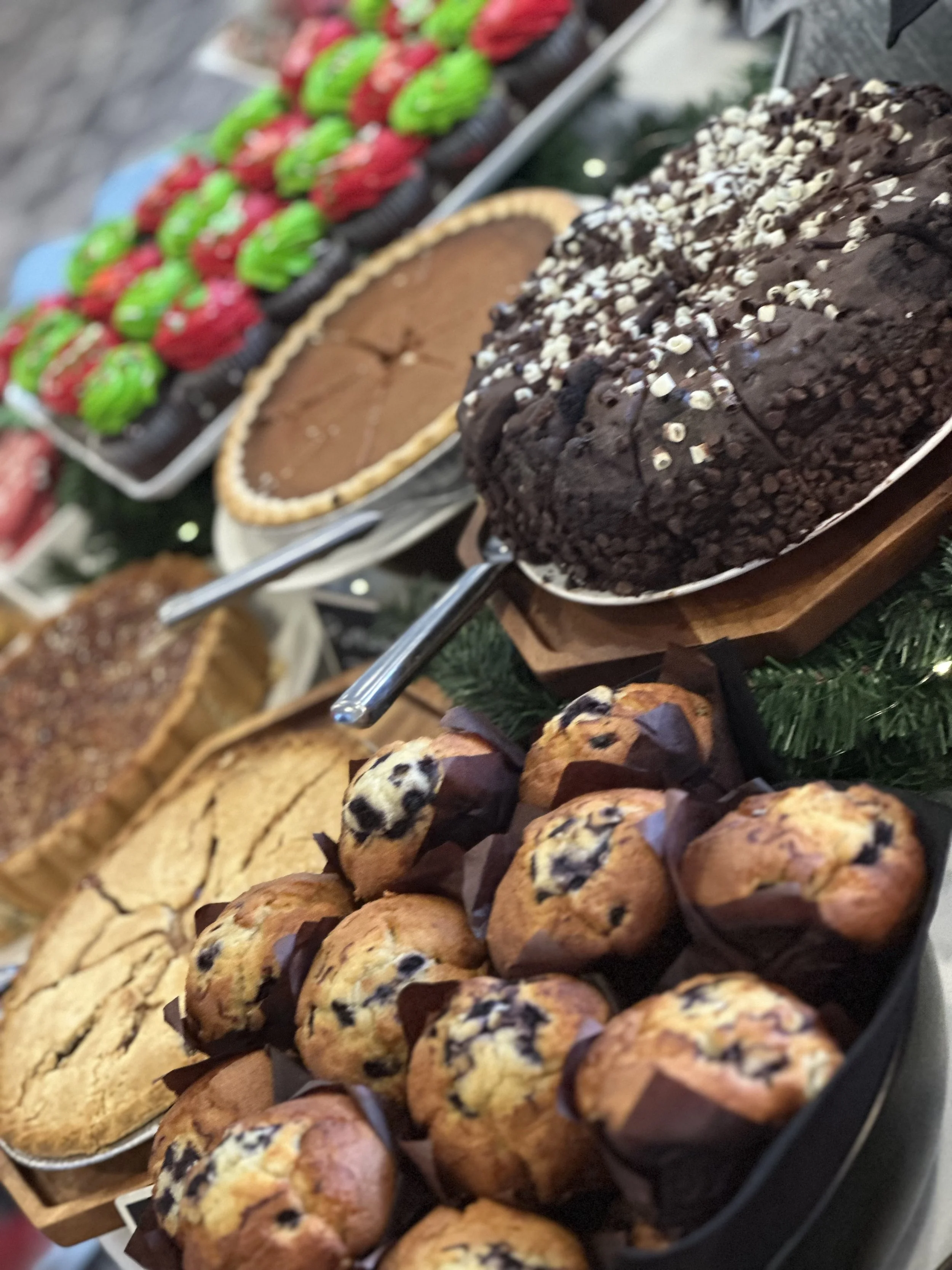Assorted baked goods including blueberries muffins, chocolate chip muffins, an Oreo cake, a chocolate pie, and multiple cupcakes, displayed on trays and plates, likely at a bakery or holiday event.