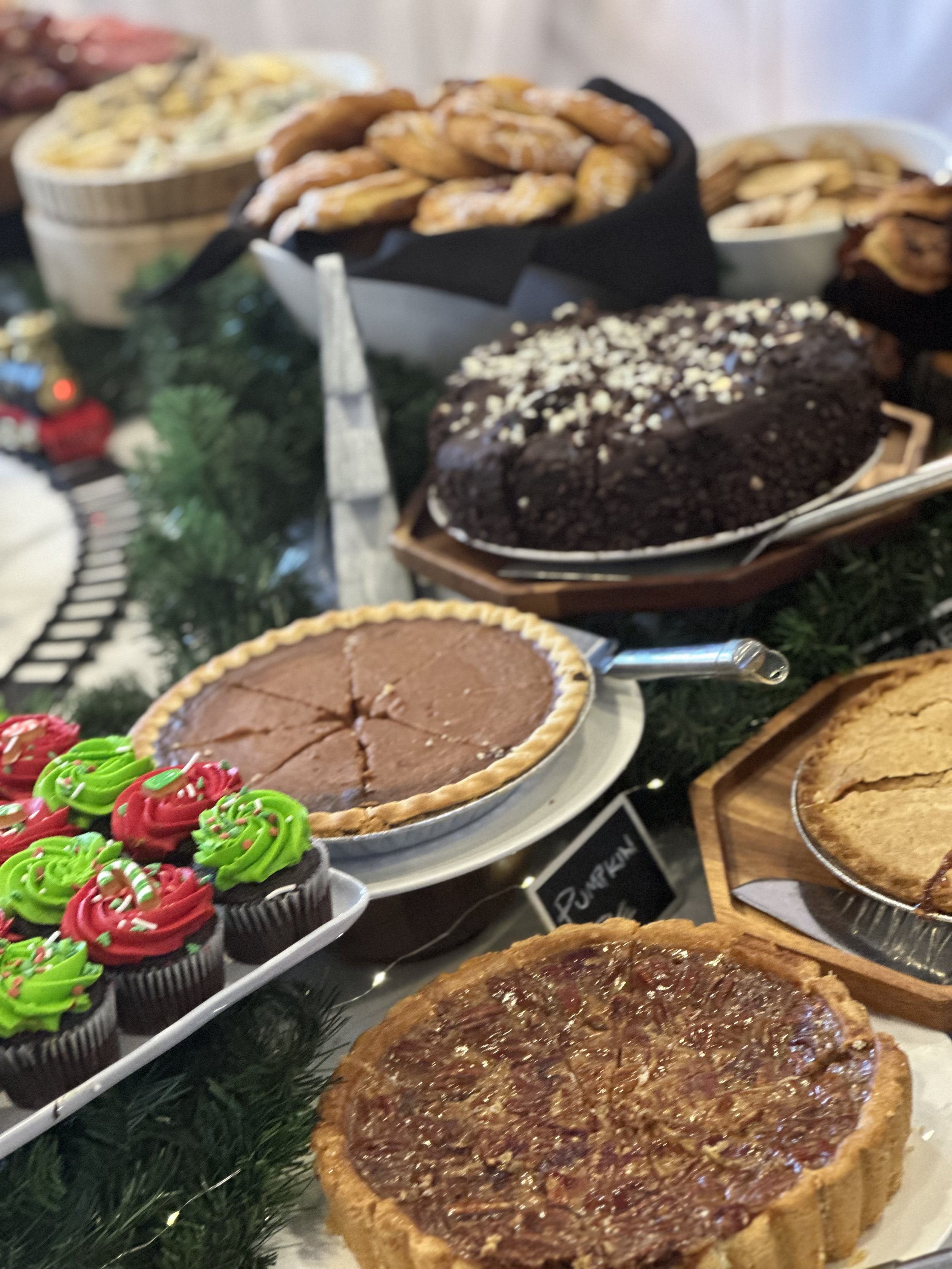 Assorted desserts on a holiday table, including cupcakes with red and green frosting, a chocolate pie, a pumpkin pie, a pecan pie, a chocolate cake with white sprinkles, and a banana pudding with vanilla wafers.