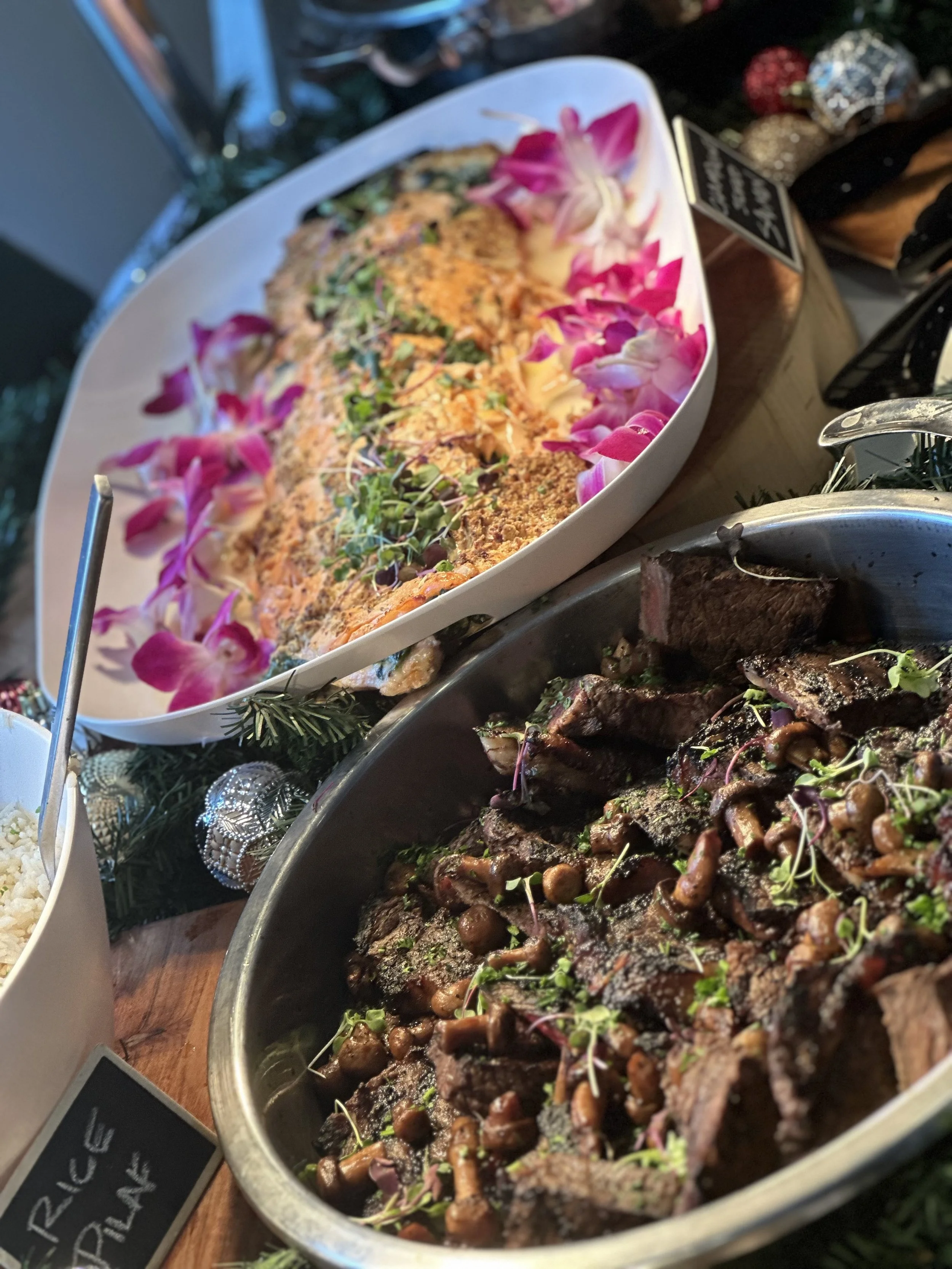 Close-up of a buffet table with sliced cooked meat garnished with microgreens, a pasta dish garnished with edible flowers, and a bowl of rice, with holiday decorations in the background.