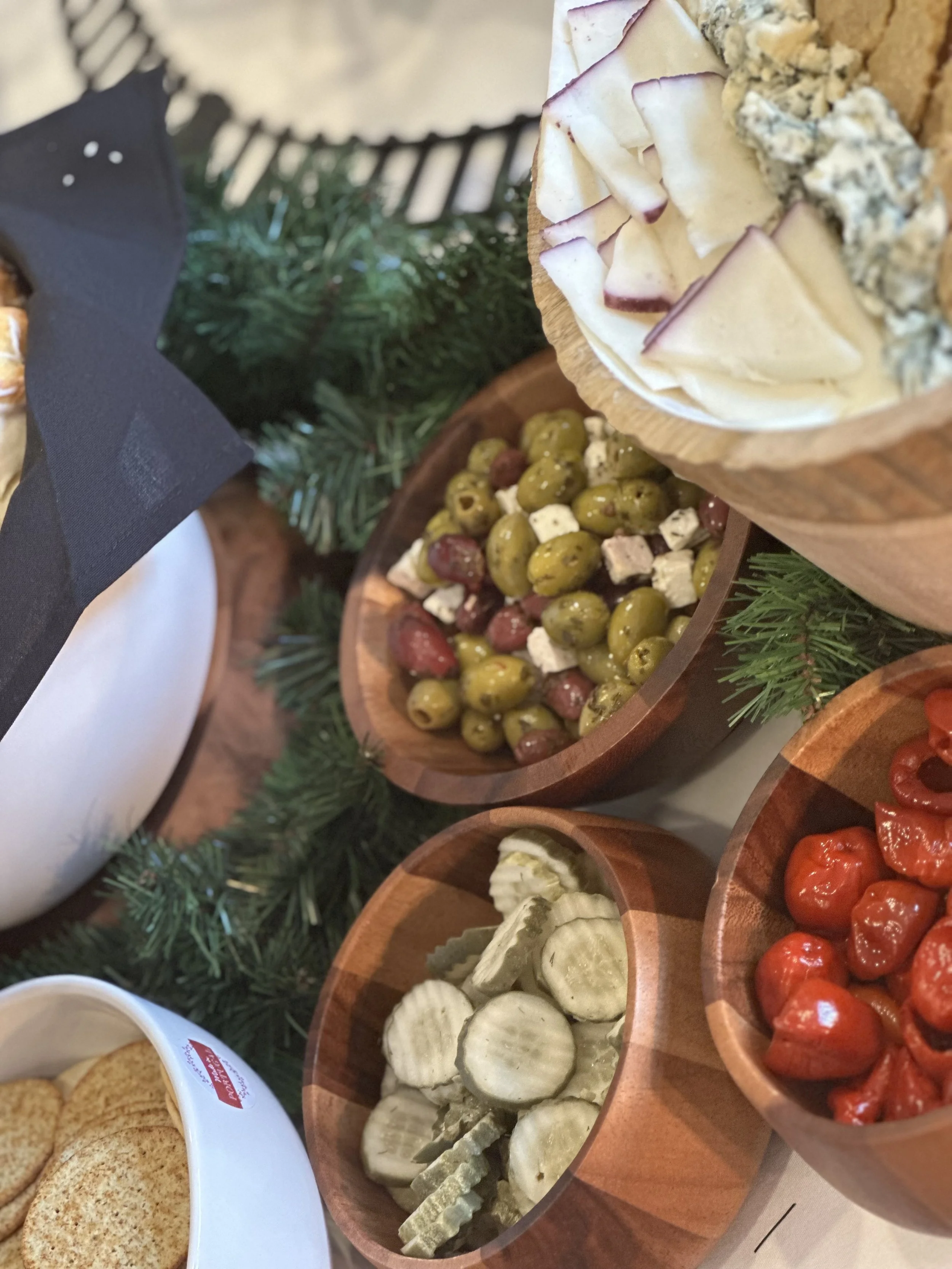 Wooden bowls filled with grapes, cheese, cherry tomatoes, and sliced cucumbers, with a plate of cookies and a decorated table with greenery.