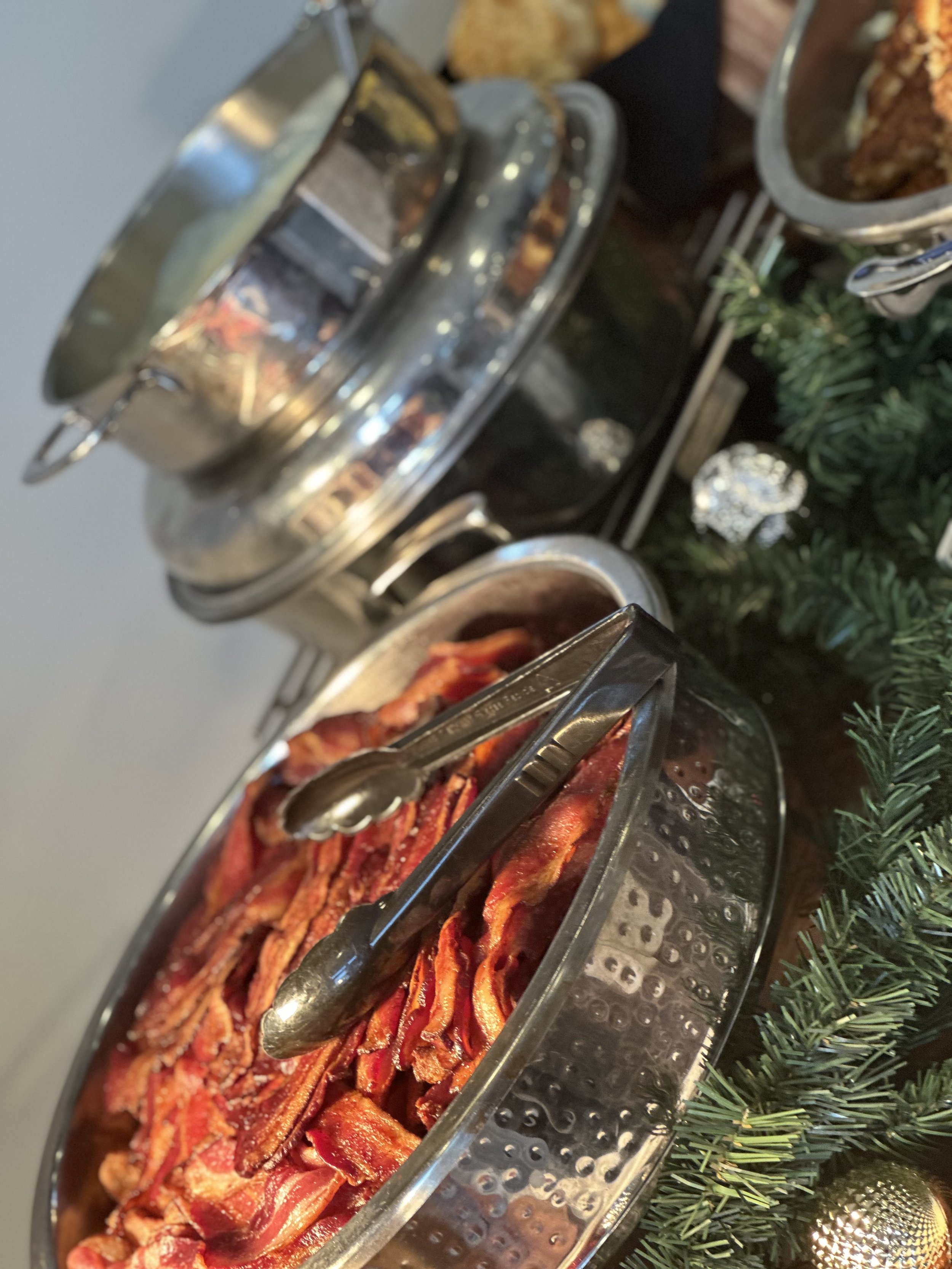 Bacon strips in a silver serving dish, with tongs resting on top, on a buffet table adorned with Christmas greenery and ornaments.