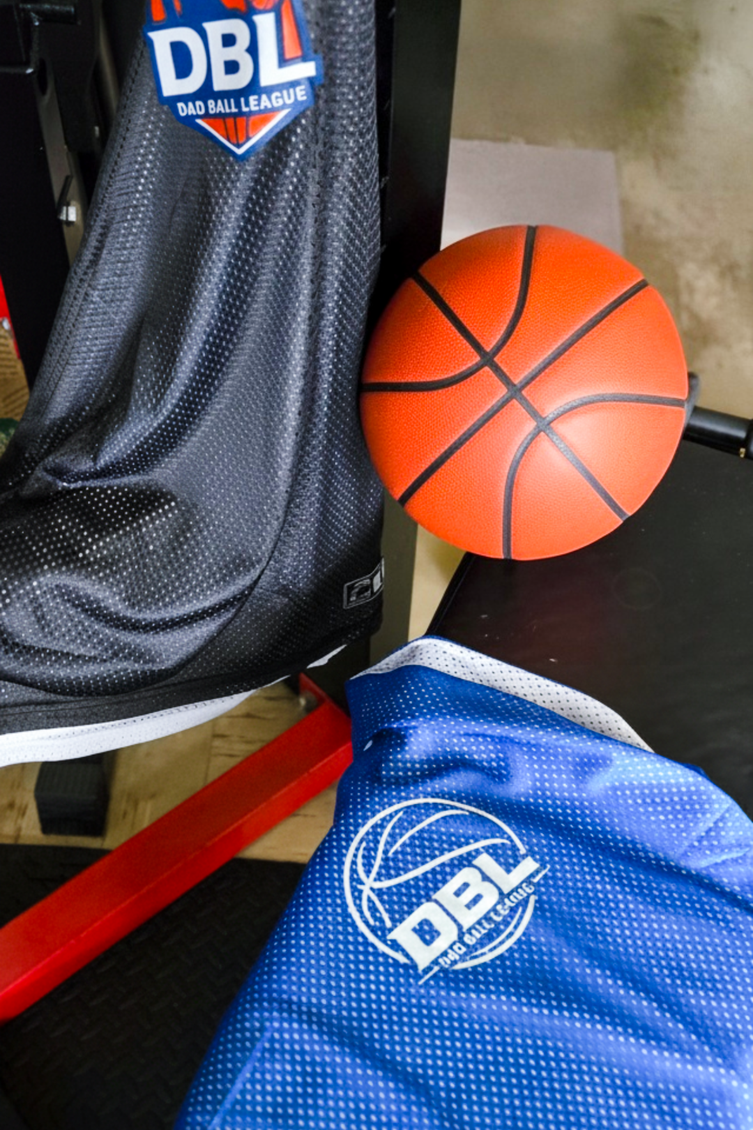 A basketball next to a black bag with the DBL (Daddy Ball League) logo on a black surface, with a blue sports jersey with the same logo partially visible in the foreground.