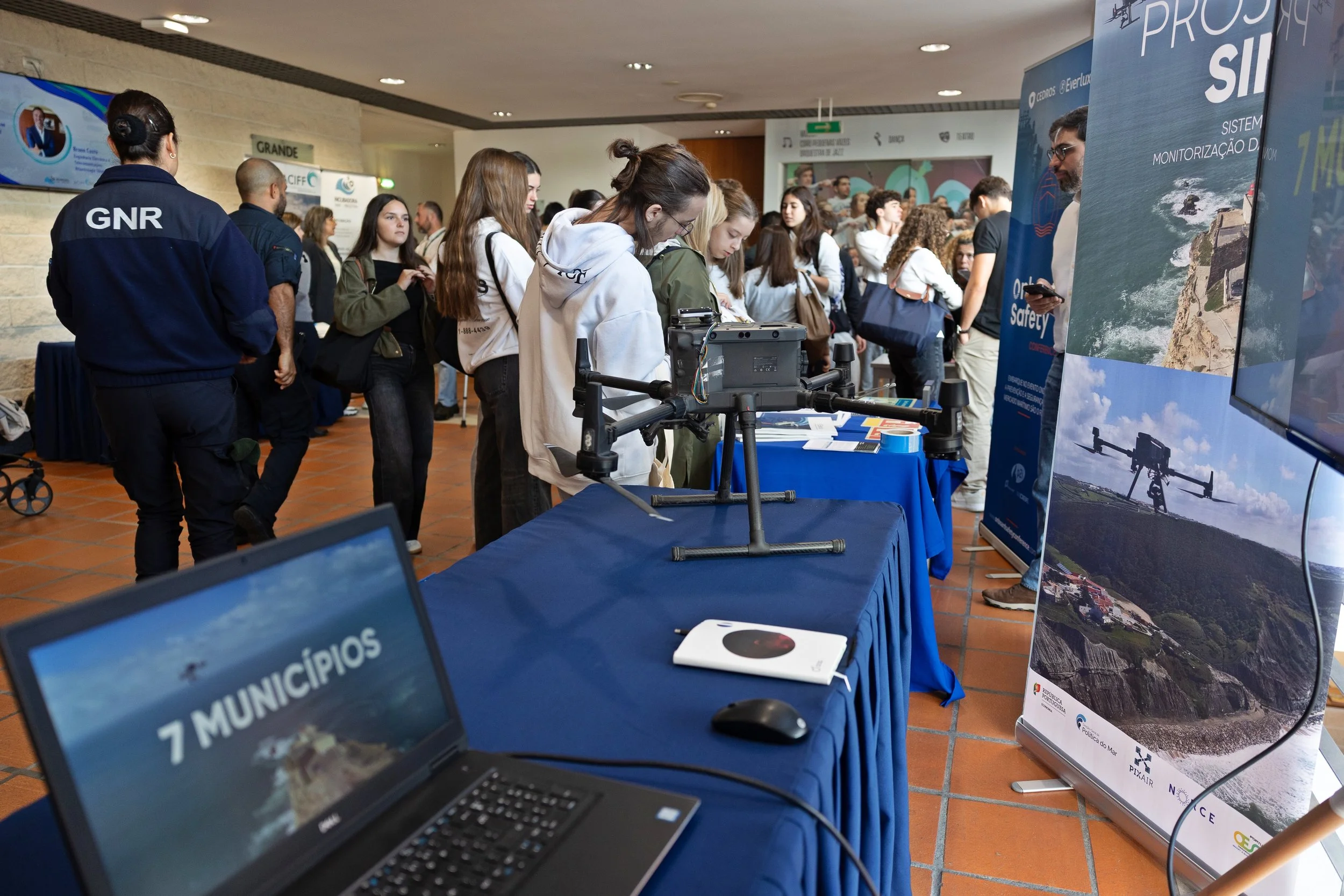 Feira com várias pessoas, expositores e banners relacionados a tecnologia e sustentabilidade, incluindo um drone em destaque na mesa.