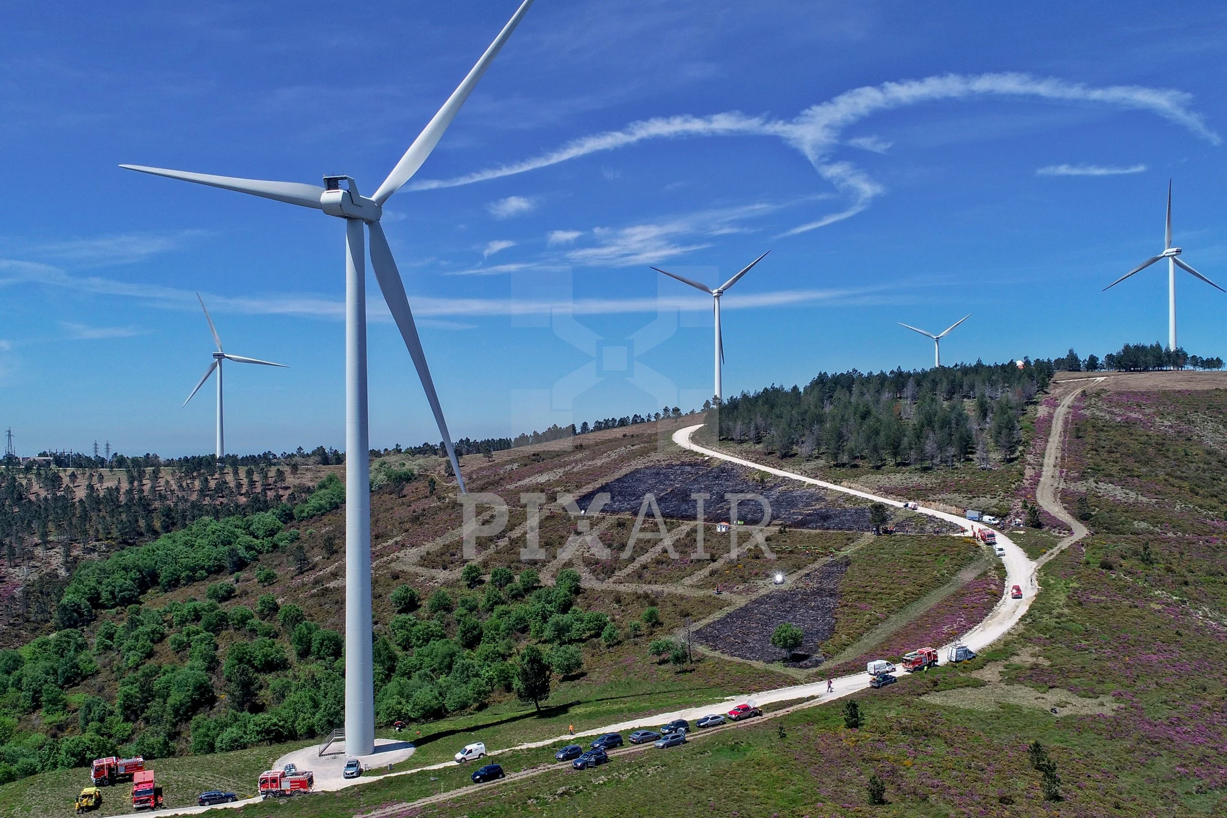 Parque de energia eólica com vários aerogeradores em uma colina, céu azul com nuvens finas e estrada com veículos.