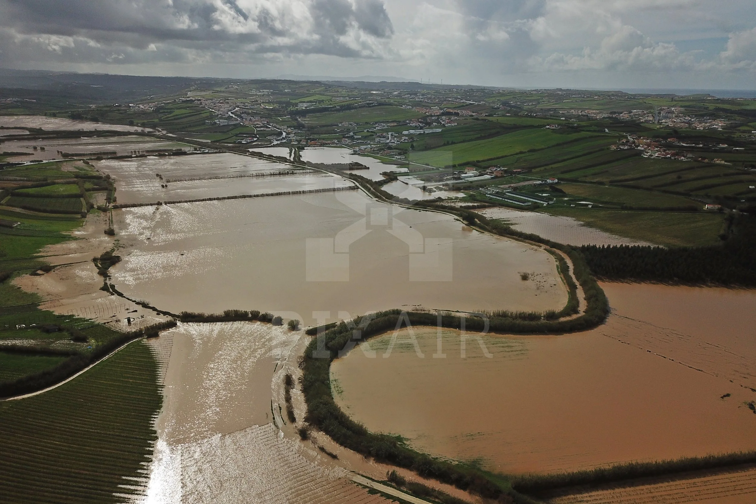 Paisagem de campos alagados com água refletindo o céu nublado, cercados por árvores e pequenas estradas.