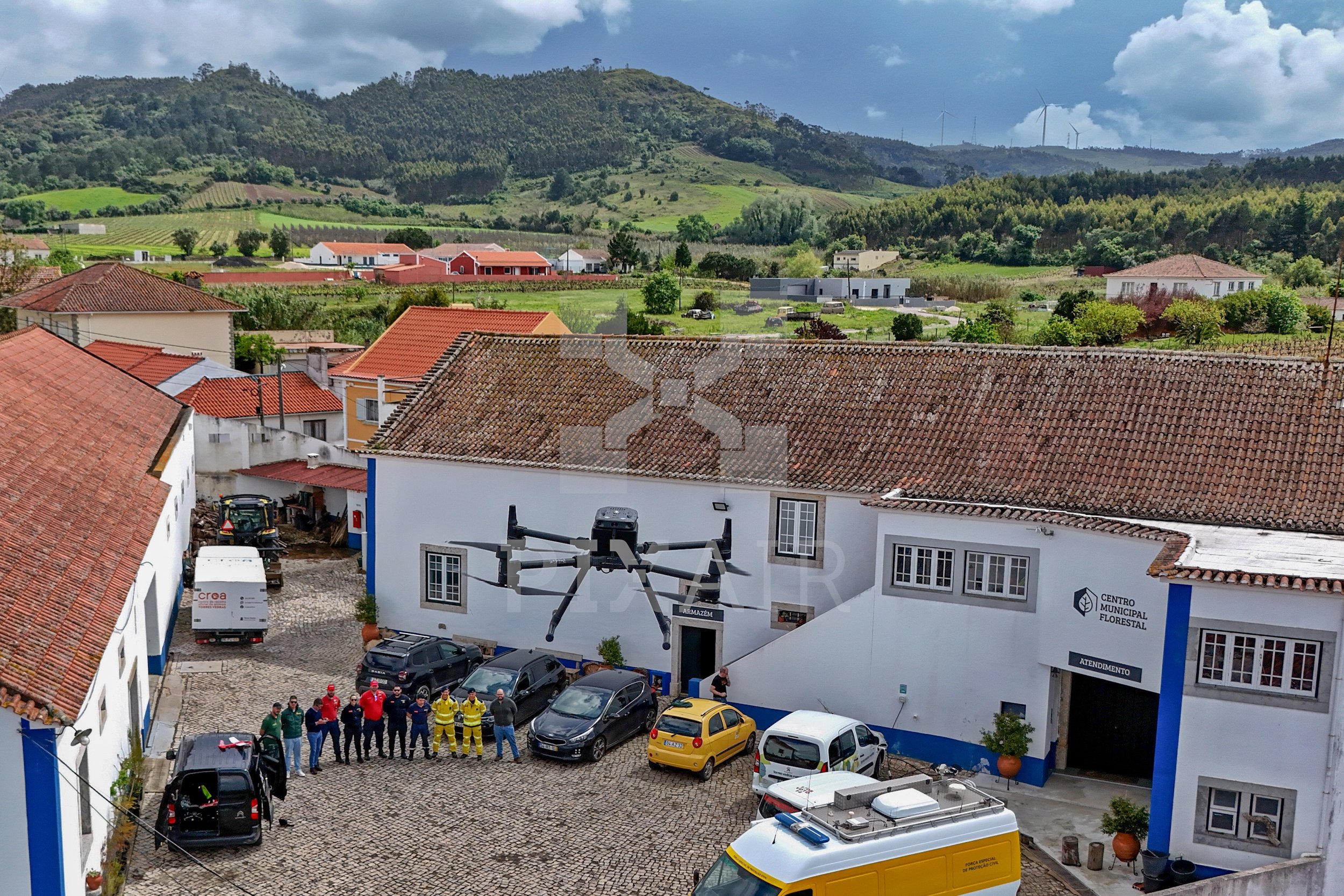Imagem de um centro comunitário com um drone gigante na parede, várias pessoas e veículos no pátio, cercado por casas e áreas verdes em uma paisagem de cidades rurais.