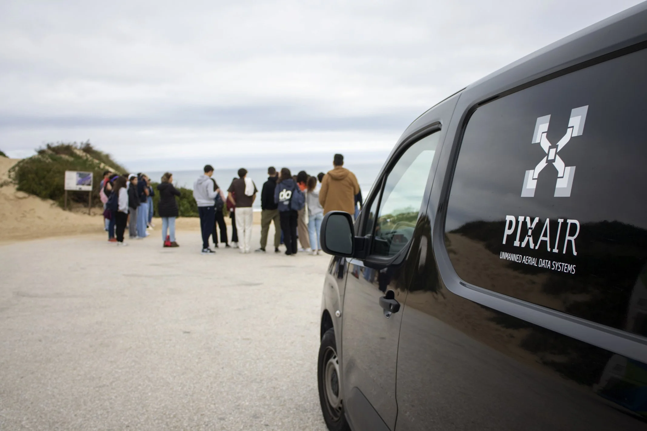 Grupo de pessoas em fila ao ar livre com um veículo estacionado ao lado, carro preto com logo da PixAIR e texto 'Unmanned Aerial Data Systems'.