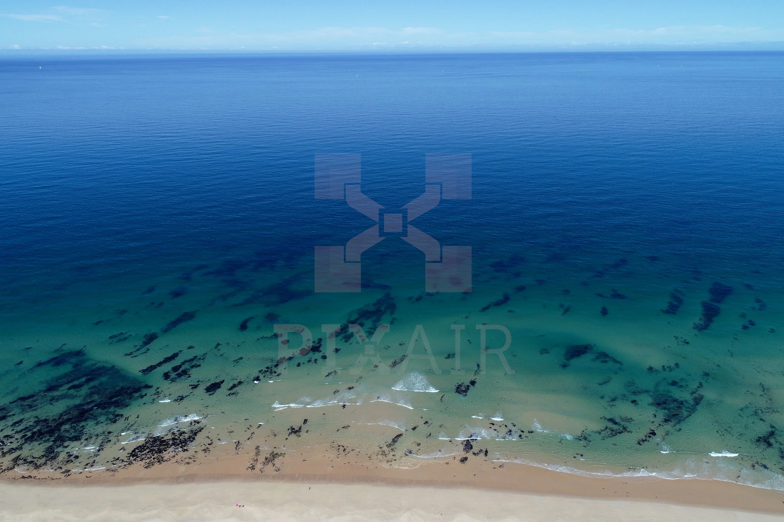 Vista aérea de uma praia com areia clara, água azul e várias manchas escuras na água, possivelmente algas ou formações submarinas, sob céu parcialmente nublado.
