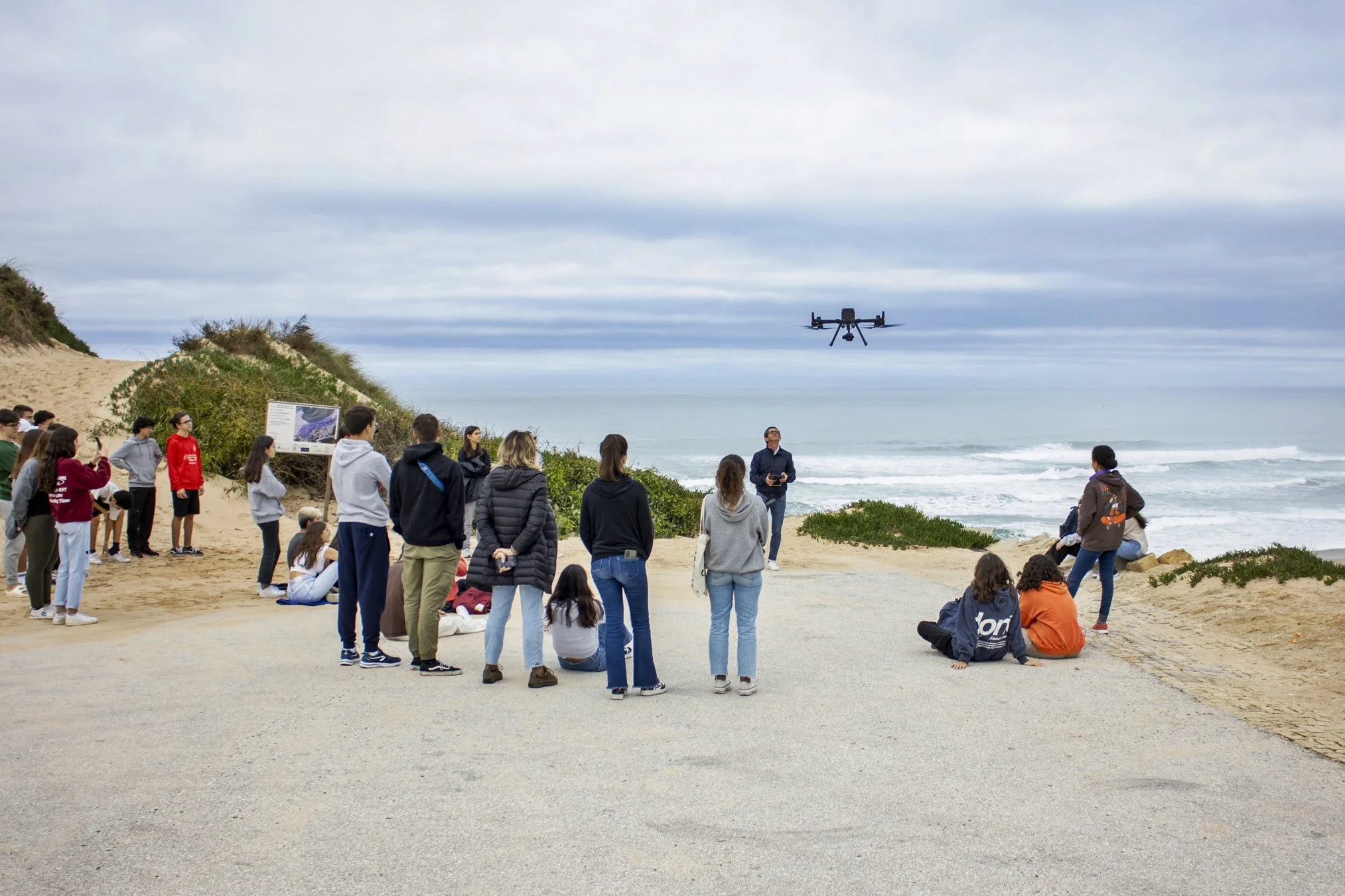 Grupo de pessoas assistindo a uma demonstração com drone na praia, com o mar e céu nublado ao fundo.