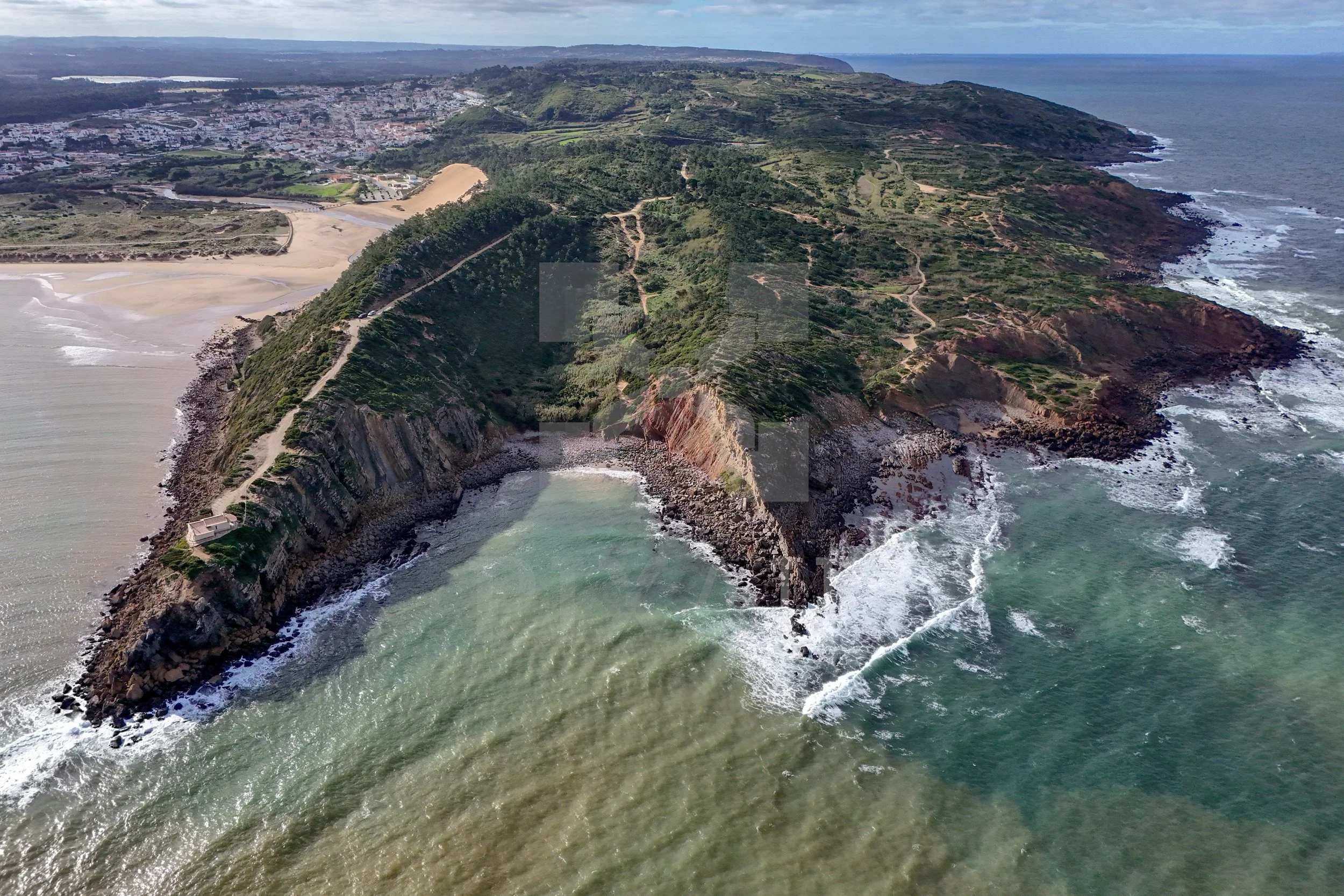 Paisagem costeira com penhasco, mar e praia na areia dourada, vista aérea de uma área com vegetação verde e trilhas na encosta, ao fundo uma cidade e o oceano com céu parcialmente nublado.