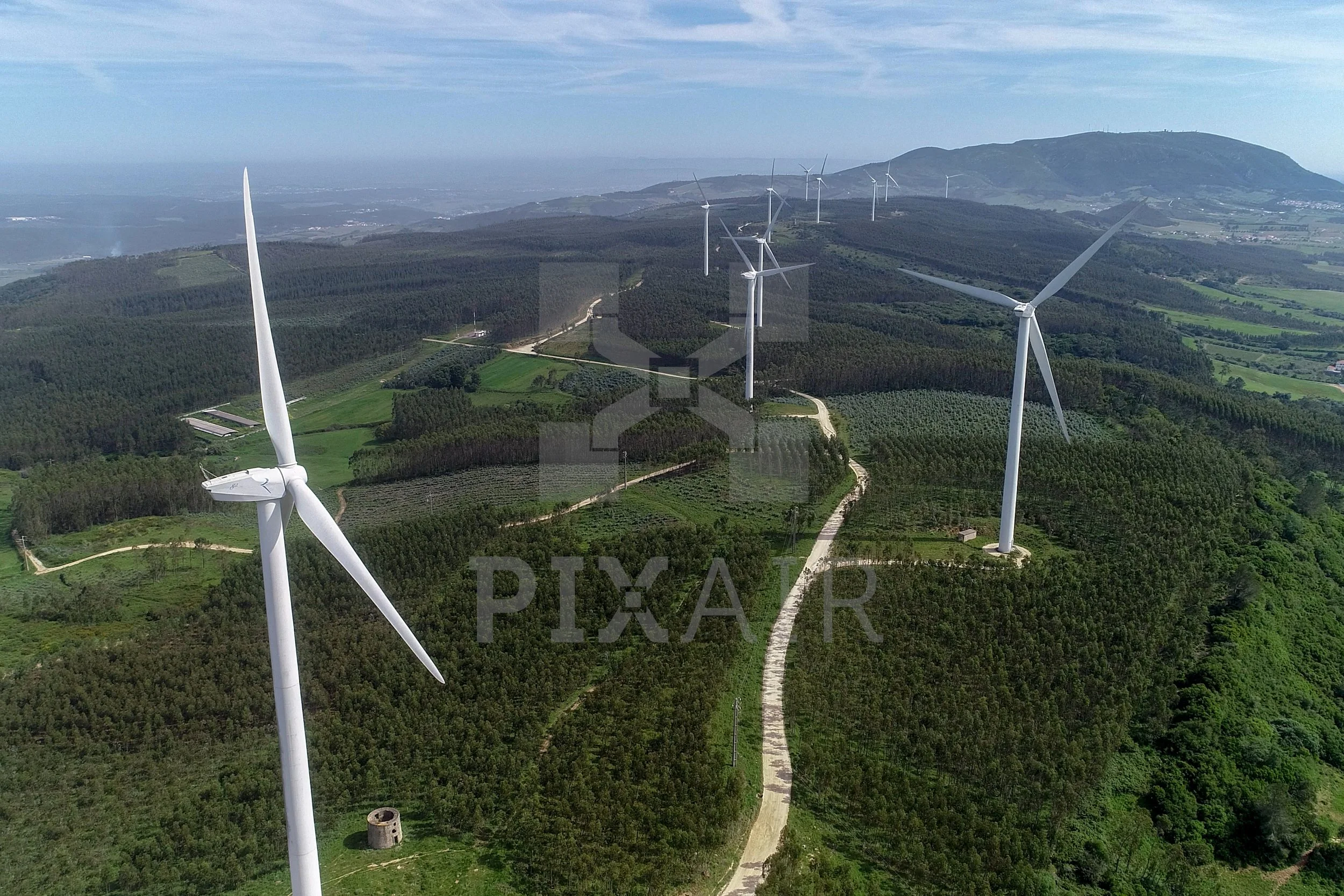 Parque eólico com várias turbinas de vento em uma paisagem de morros verdes com estrada de terra e árvores ao redor.