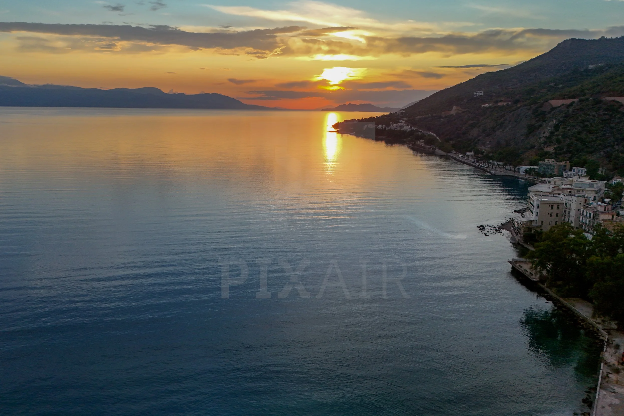 Pôr do sol sobre o mar, com montanhas ao fundo e prédios à direita na orla.
