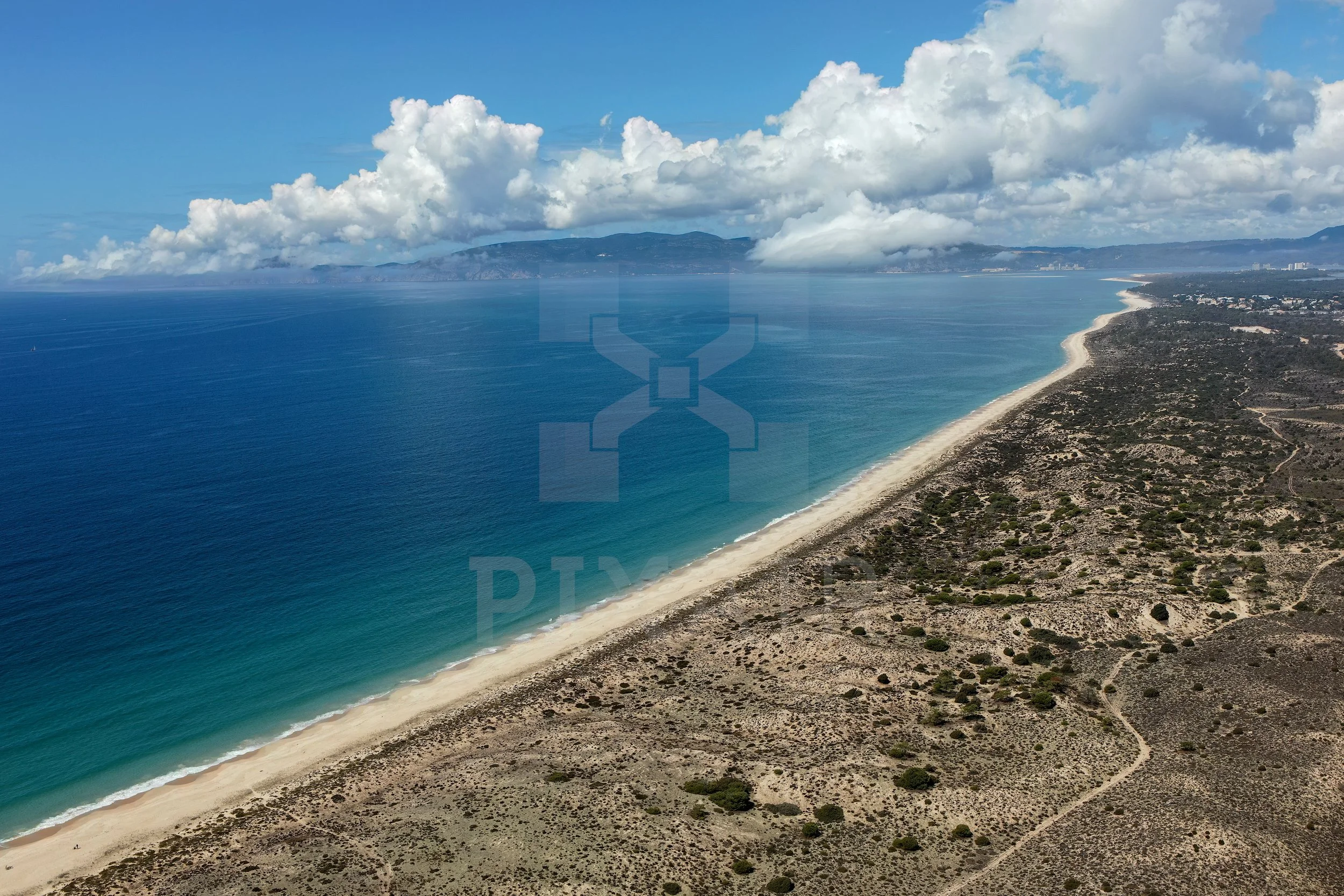 Vista aérea de uma praia extensa com areia clara, águas azuis e marés tranquilas, acompanhada por uma costa rochosa e vegetação dispersa, com nuvens brancas no céu