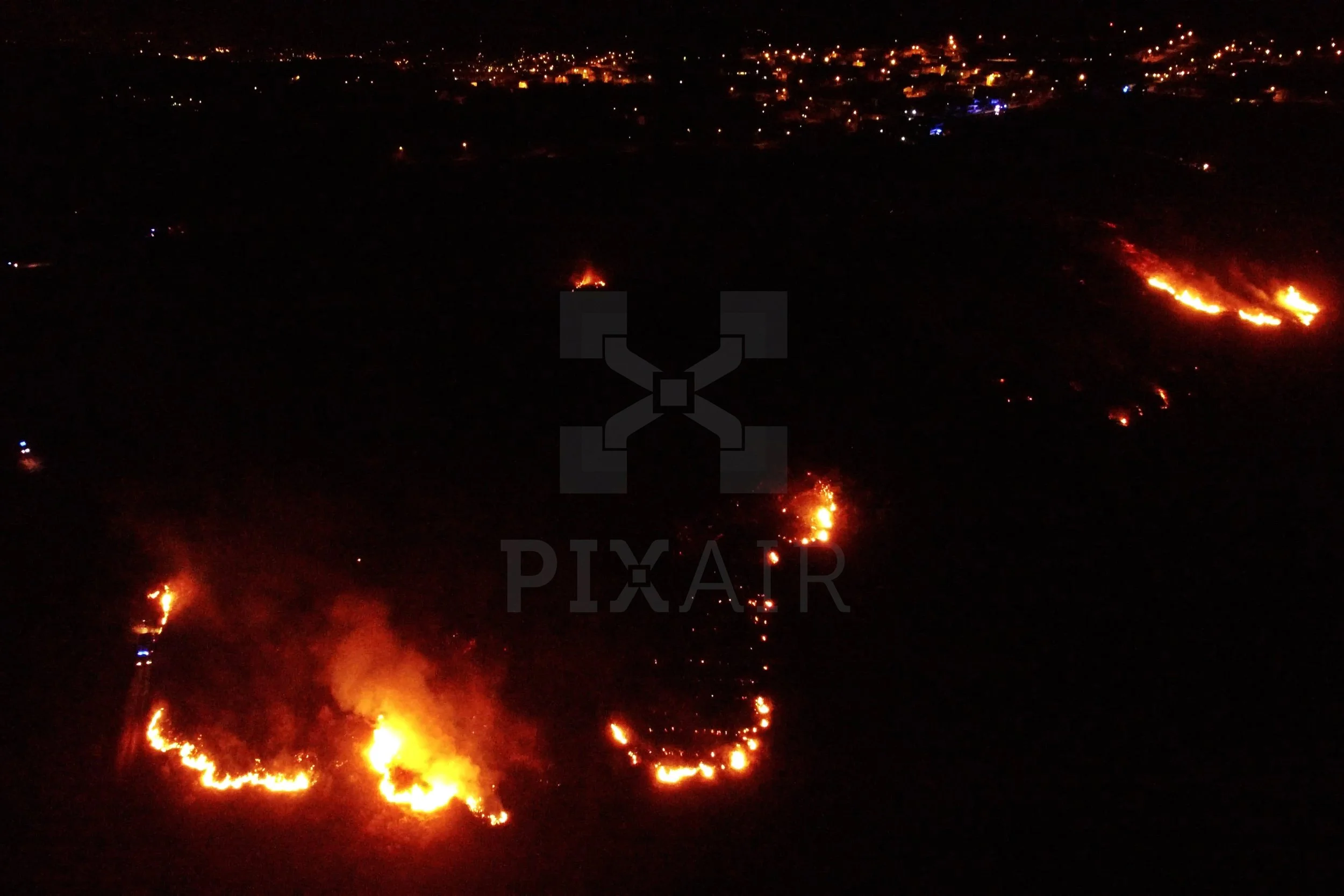 Fogo de incêndio em uma área aberta à noite, com várias chamas visíveis e fumaça na vegetação.