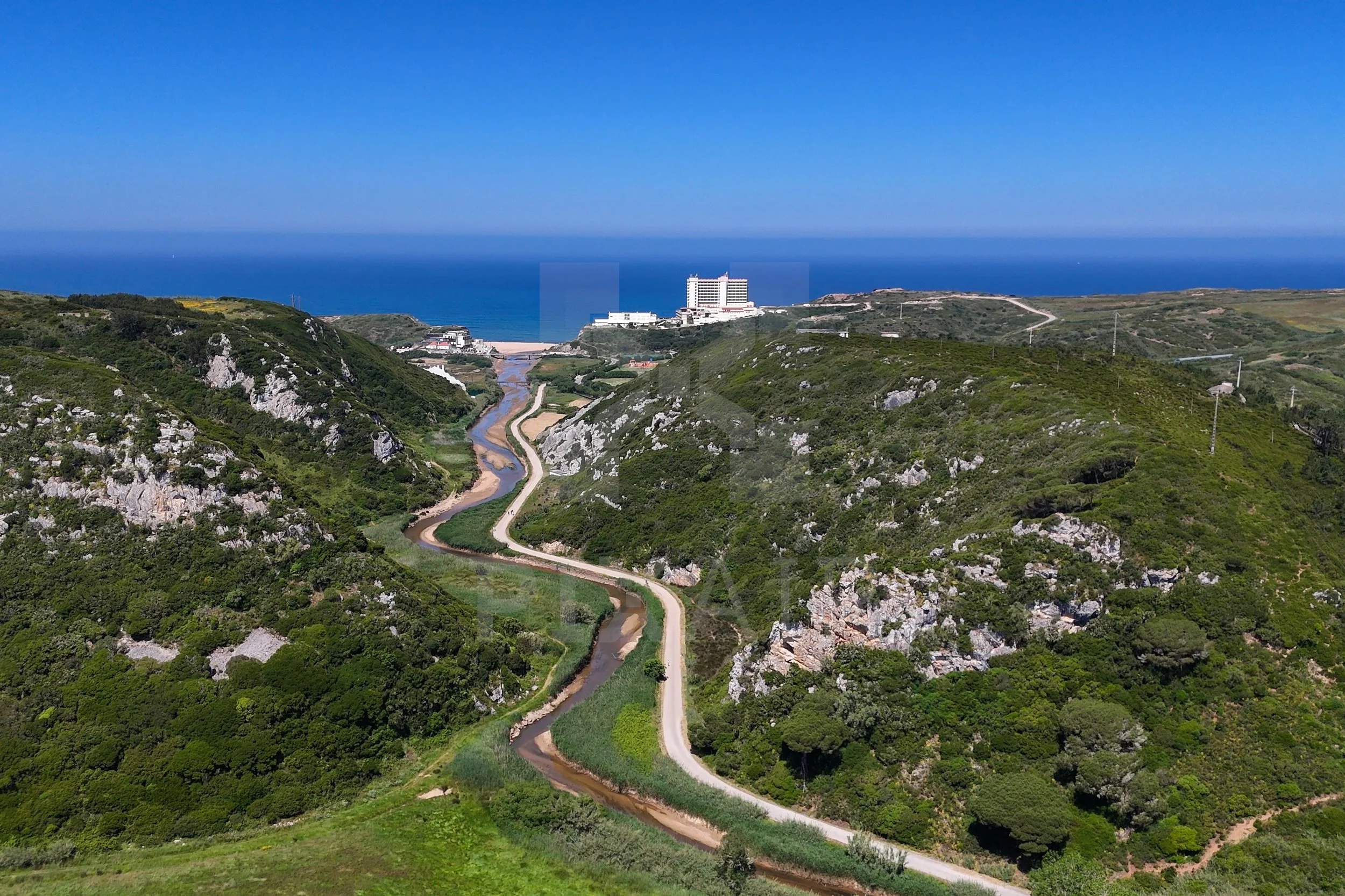 Paisagem com rio serpenteante passando por colinas verdes, com edifícios ao fundo e mar ao horizonte.