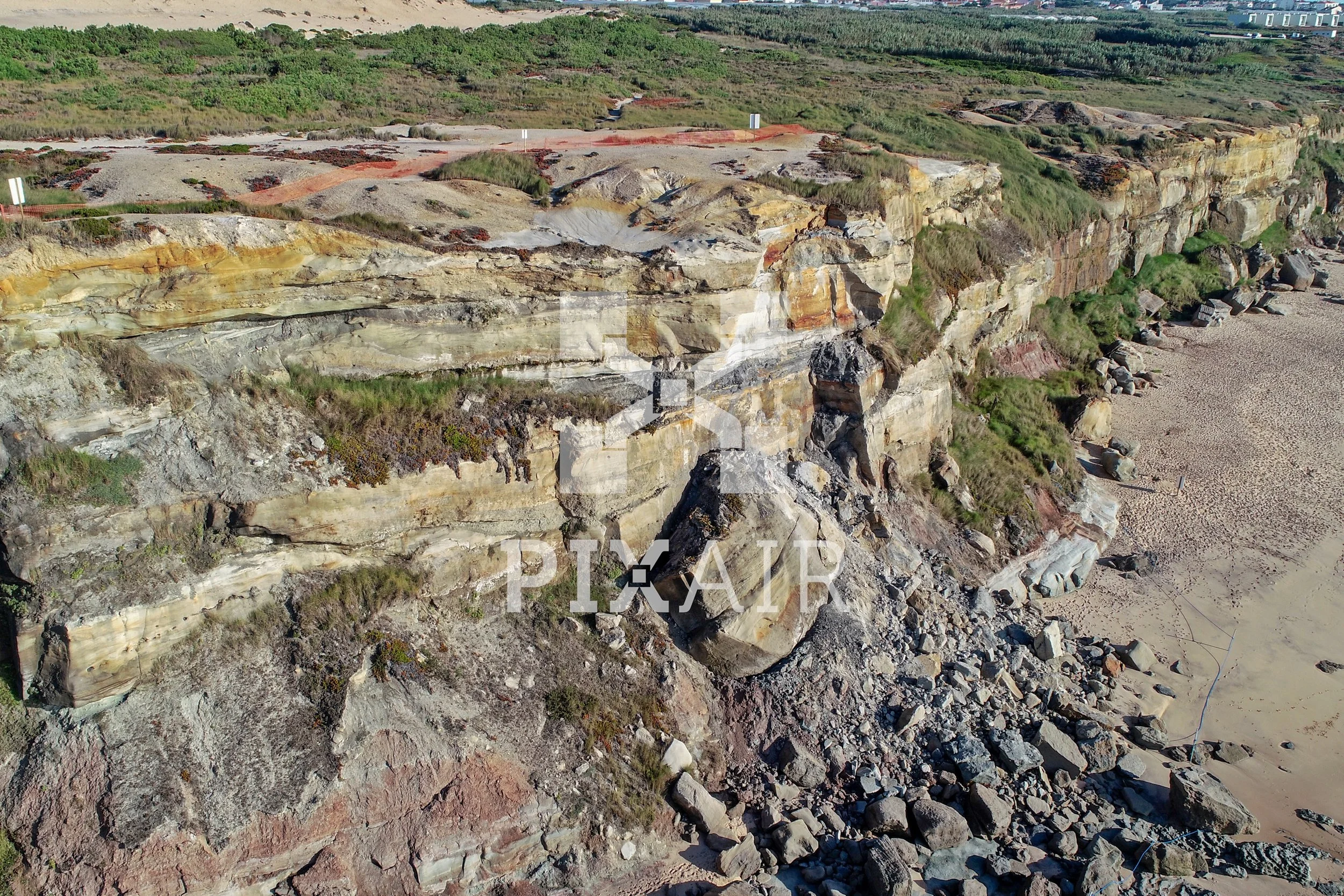 Imagem de uma formação de falésia de rochas coloridas junto ao mar, com vegetação ao fundo e uma praia de areia na base.